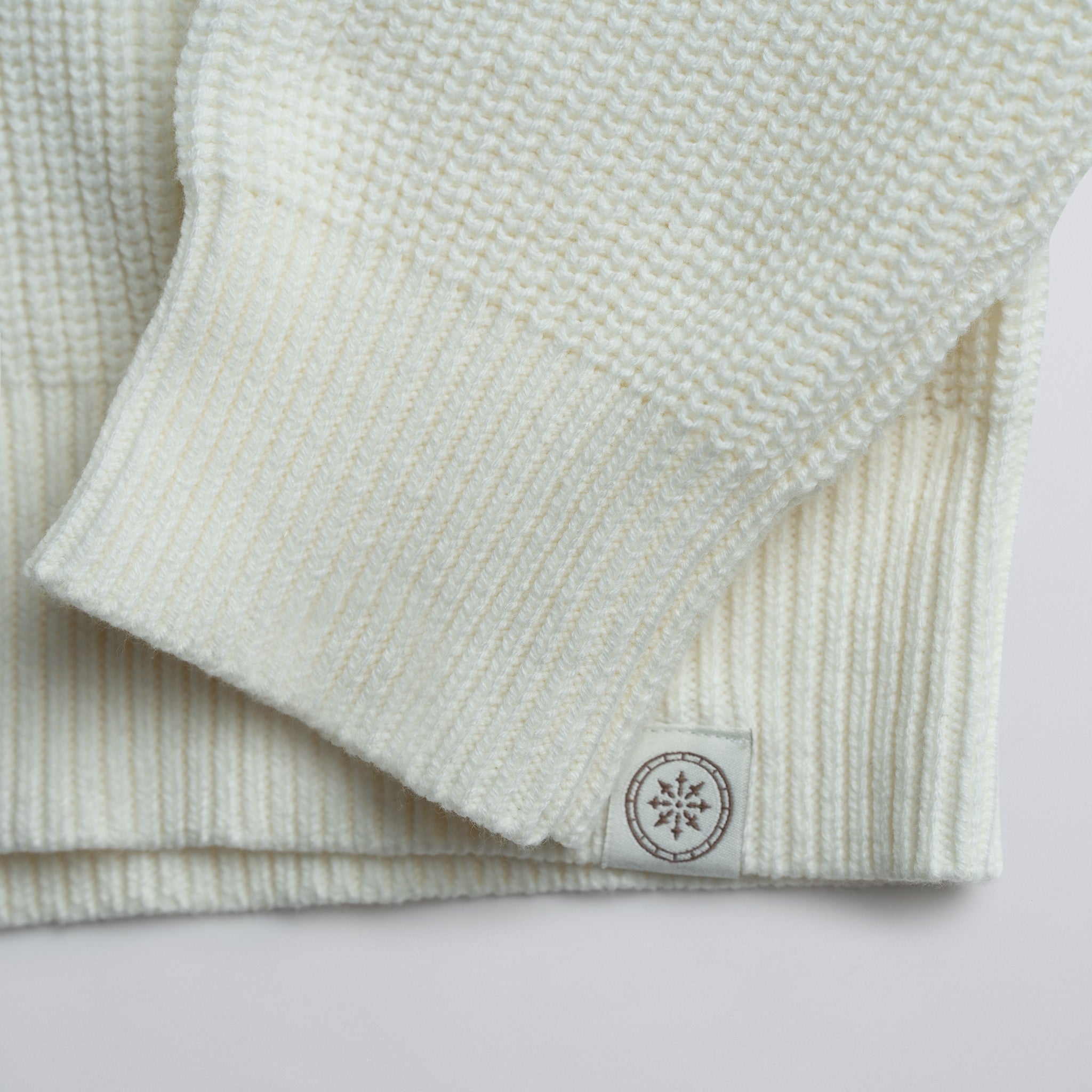 Close-up of a folded cream sweater with a logo on a plain background.