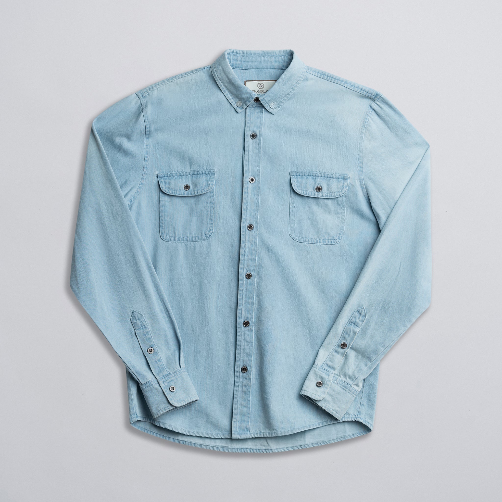 A blue denim shirt with two front pockets, button-up front, and long sleeves laid out flat on a white background.
