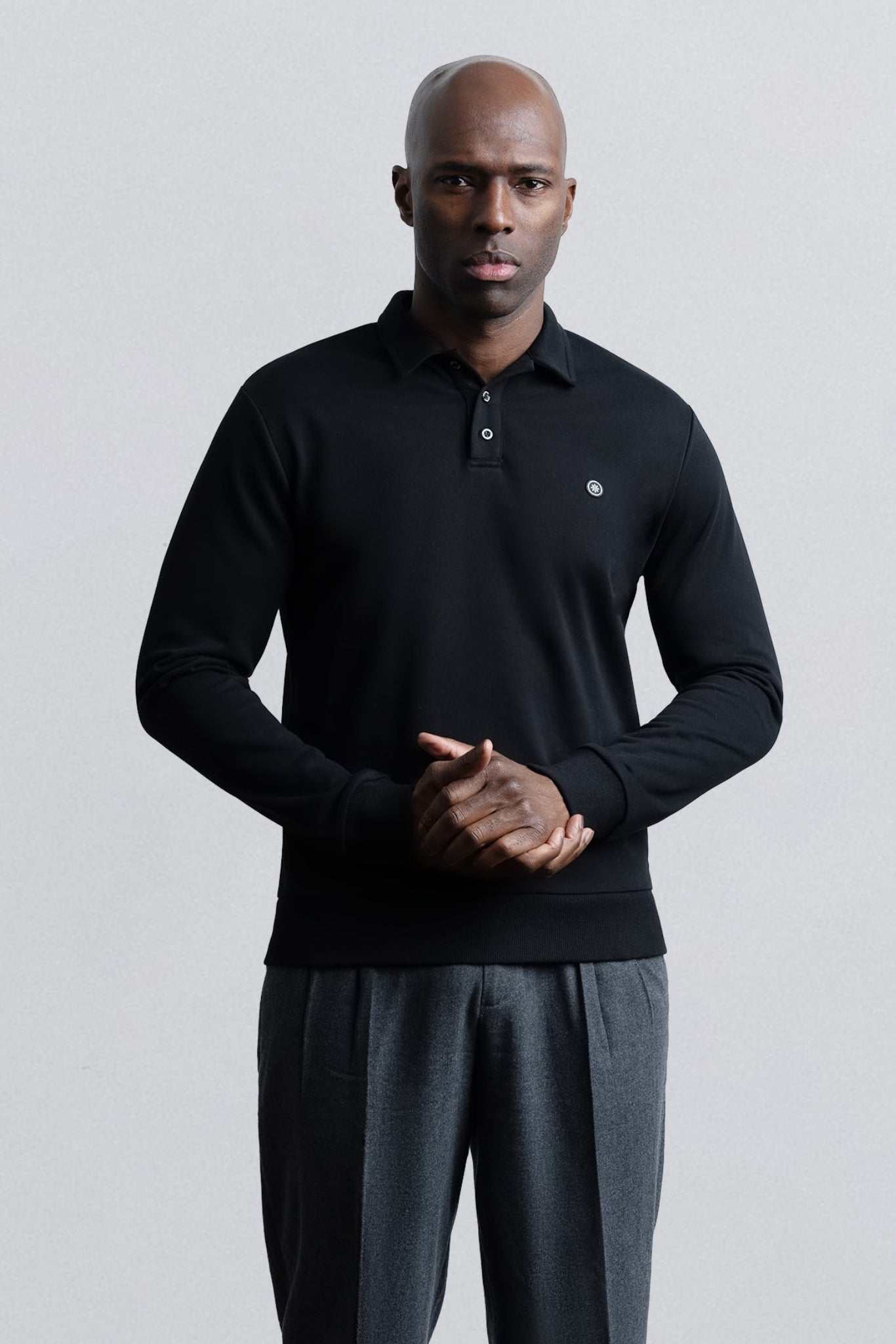 A man wearing a black long-sleeve polo shirt with a small embroidered logo on the chest, paired with dark gray trousers, standing against a light gray studio background.