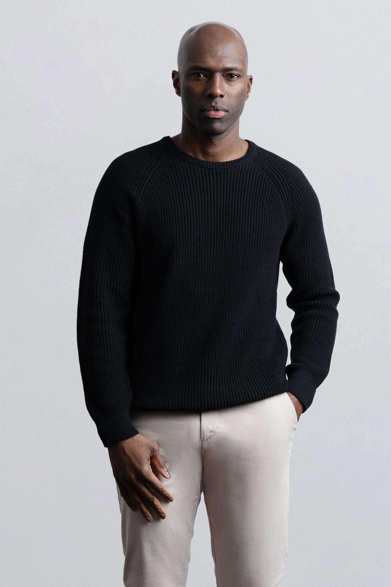 Man wearing a black sweater and light-colored pants against a plain background