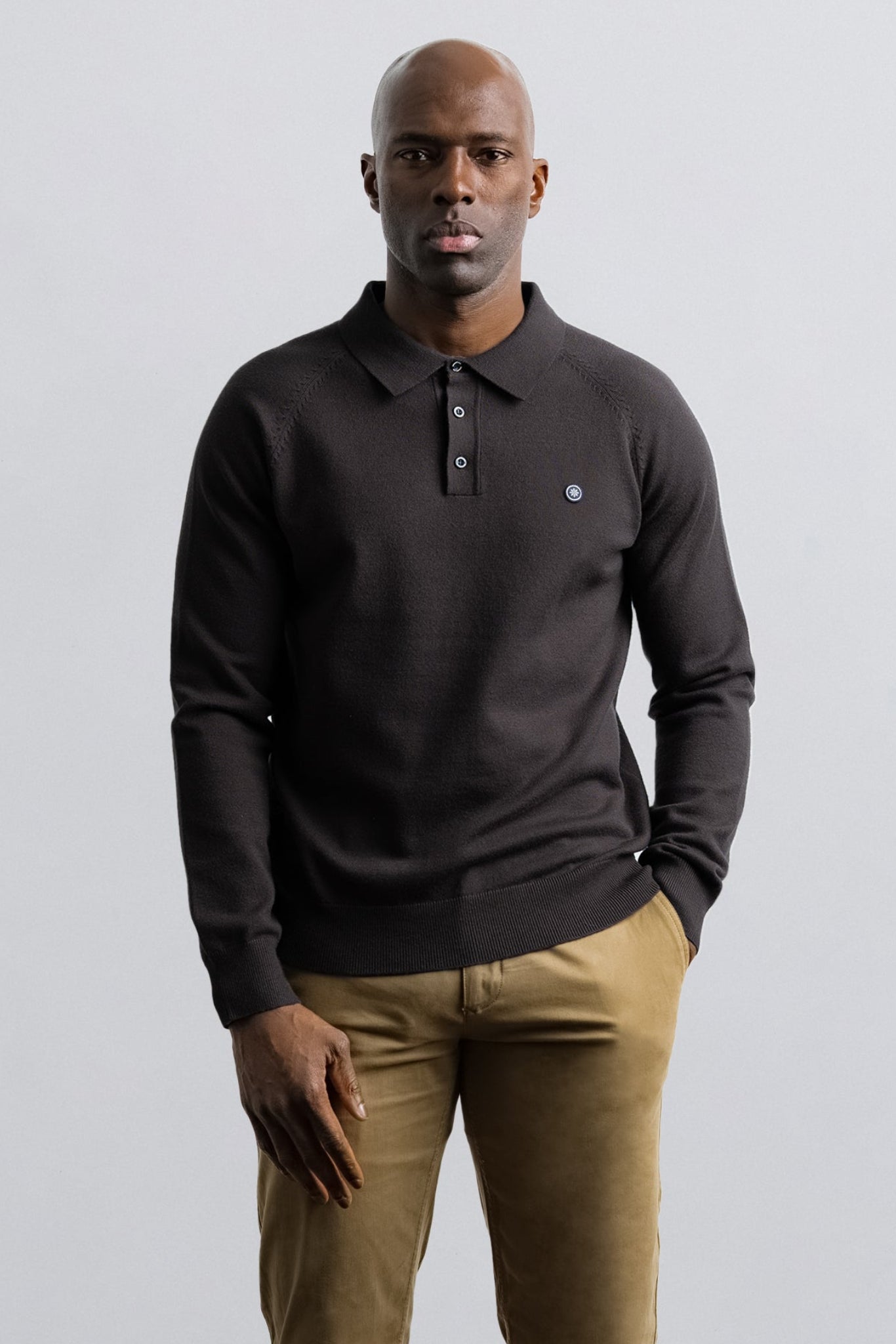 A man wearing a dark charcoal gray long-sleeve knit polo sweater and khaki trousers, standing against a light gray studio background.