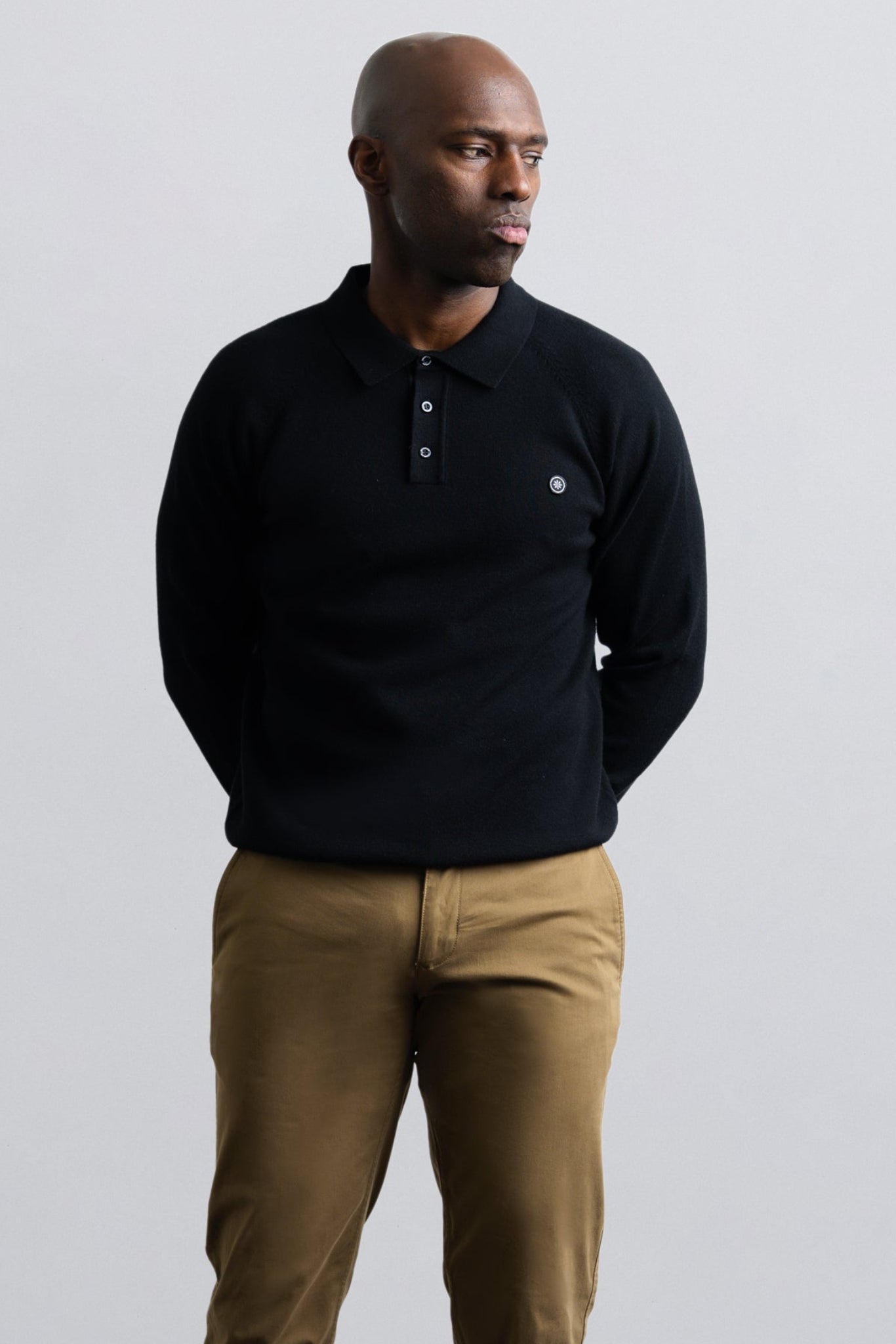 A man wearing a black long-sleeve polo shirt with a small white embroidered logo on the chest, paired with tan pants, standing against a light gray studio background.