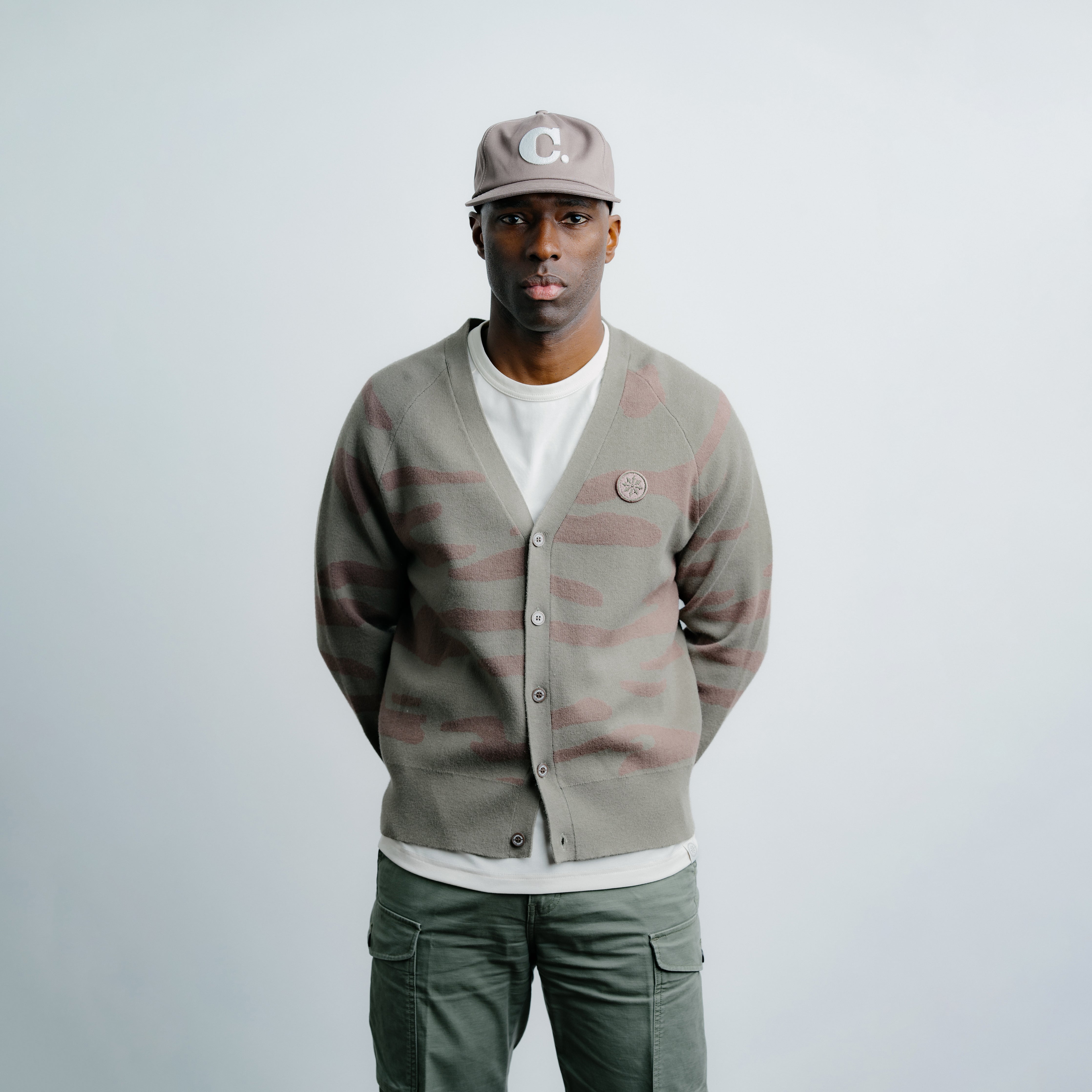 Man wearing a camouflage cardigan, white shirt, and green pants on a light gray background