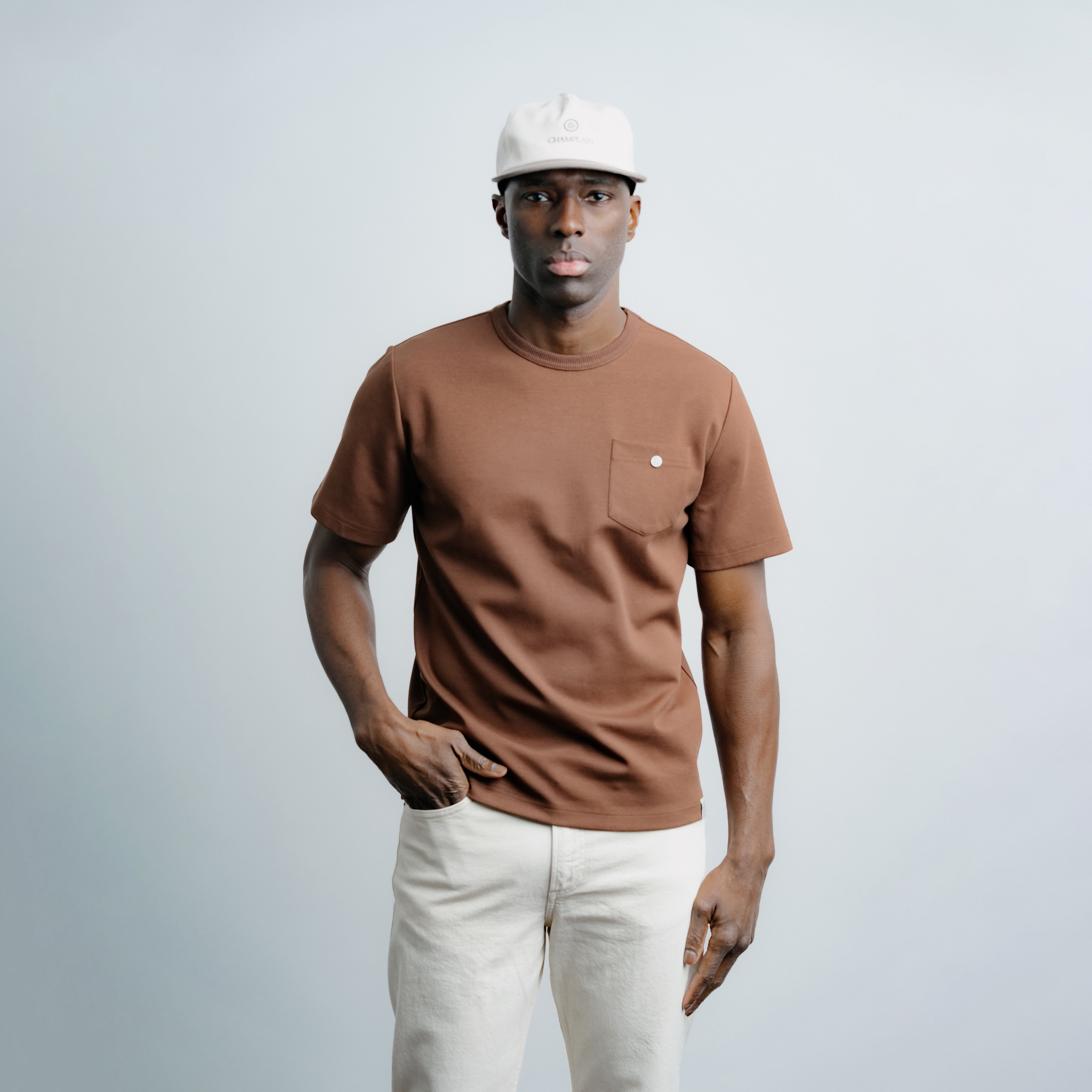 Man wearing a brown t-shirt with a pocket and white pants against a light gray background