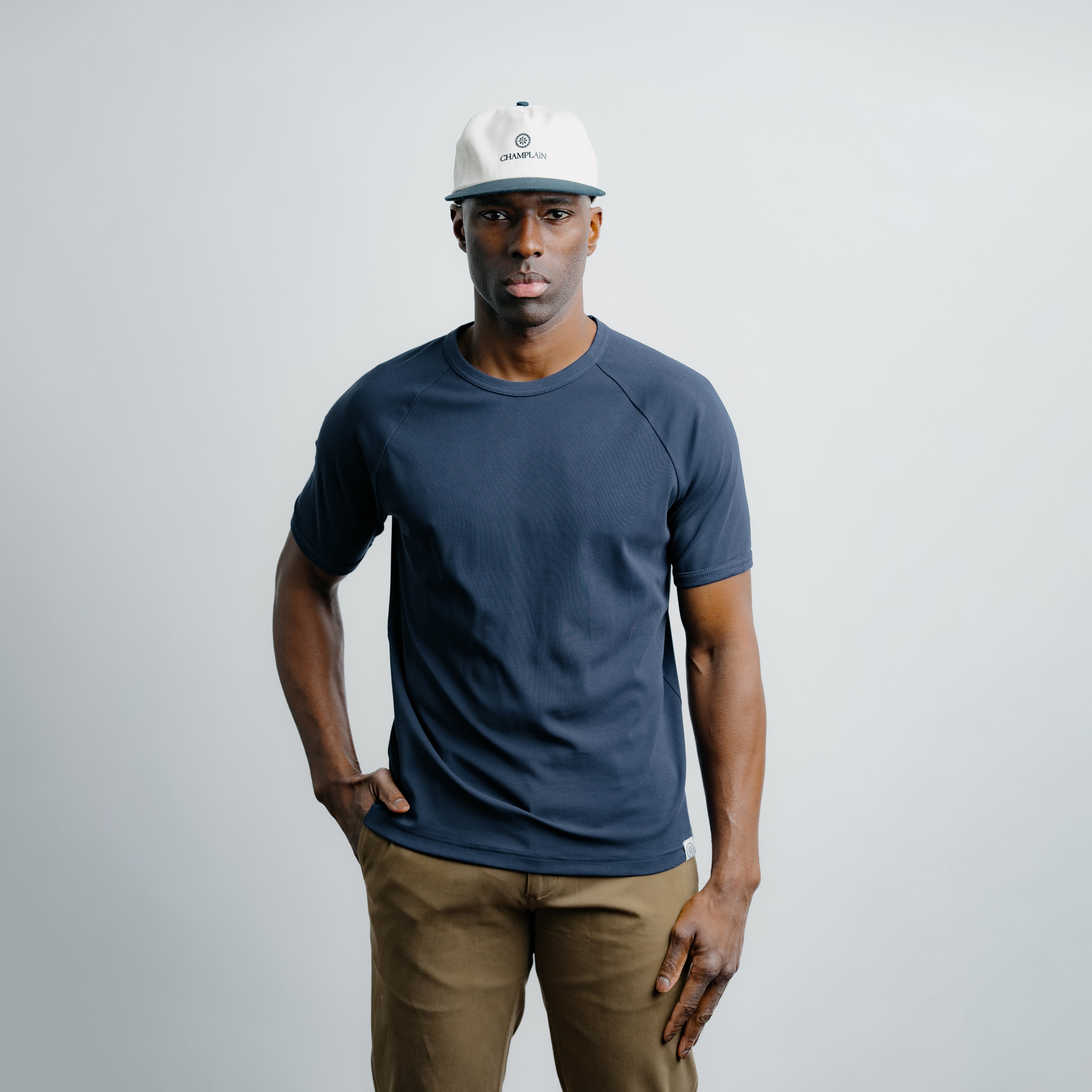 Man wearing a navy t-shirt and beige pants with a white cap on a gray background