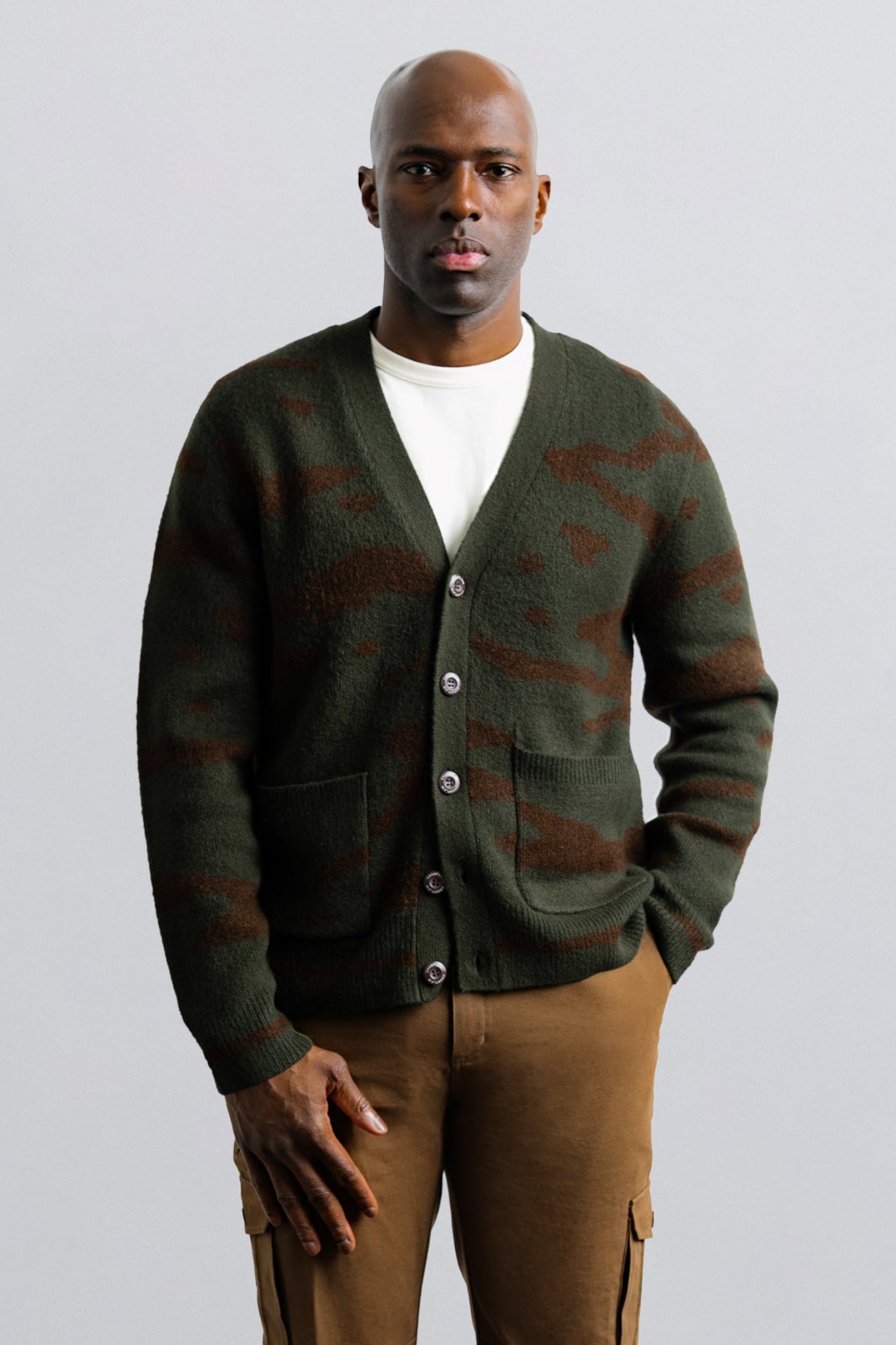 Man wearing a green cardigan over a white shirt and brown pants on a light gray background