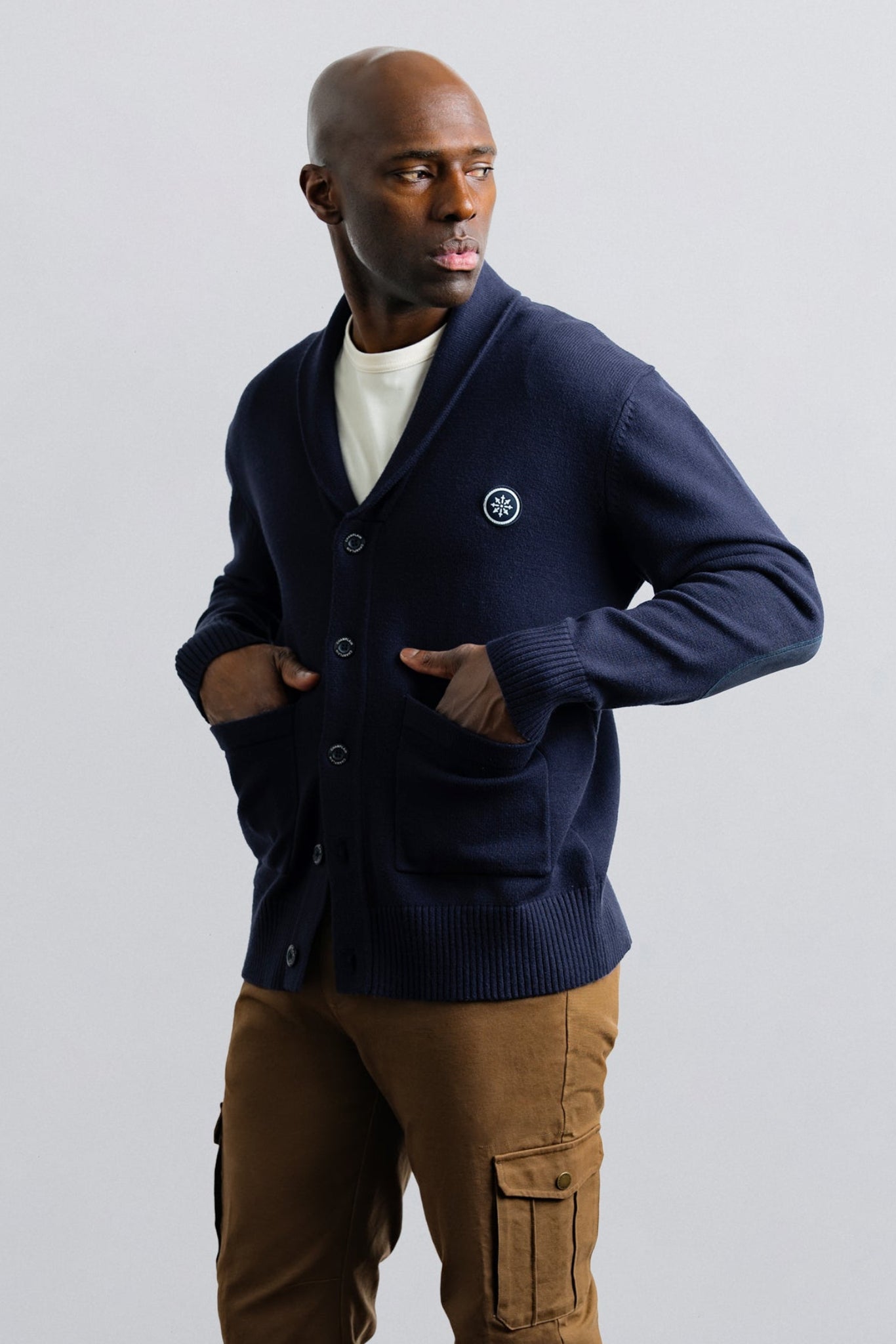 Man wearing a navy cardigan over a white shirt with brown pants on a light gray background