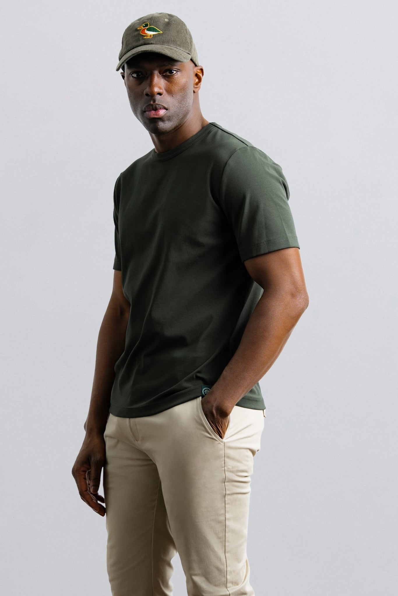 Man wearing a green t-shirt, beige pants, and a cap on a light gray background
