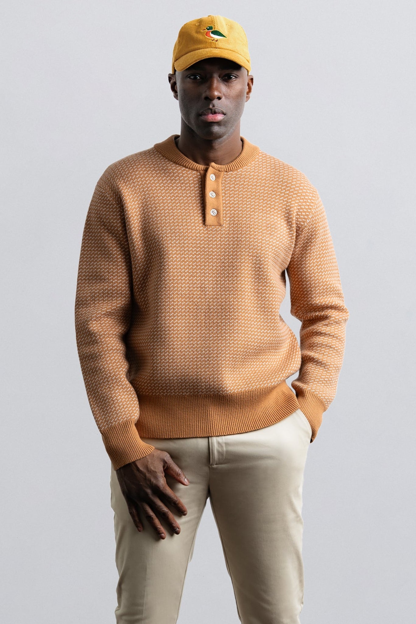 Man wearing a brown sweater and beige pants with a yellow cap on a light gray background