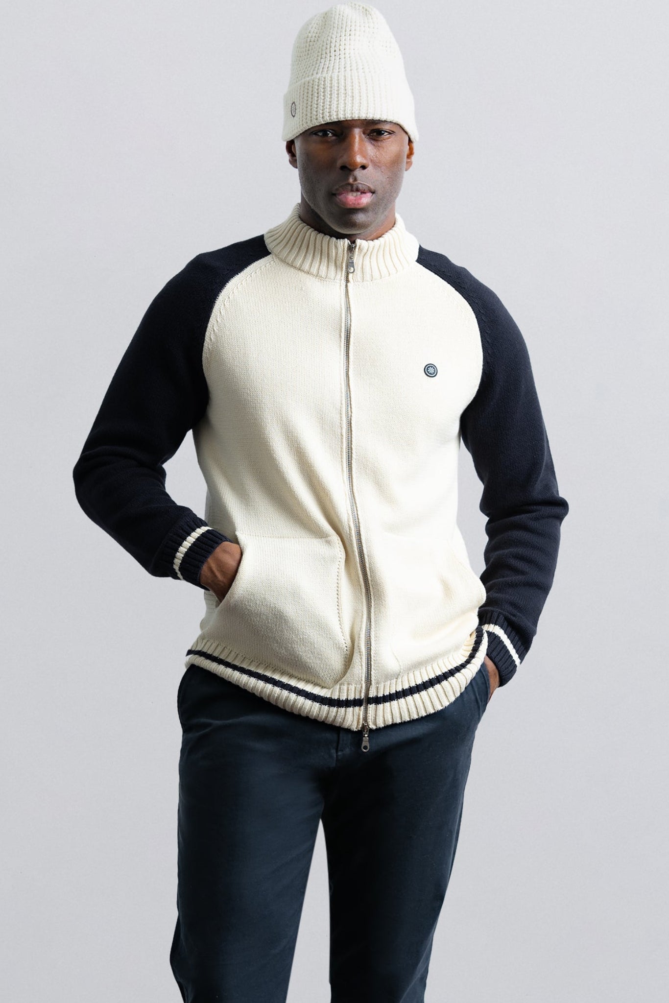 Man wearing a cream and navy jacket with a white beanie on a light gray background