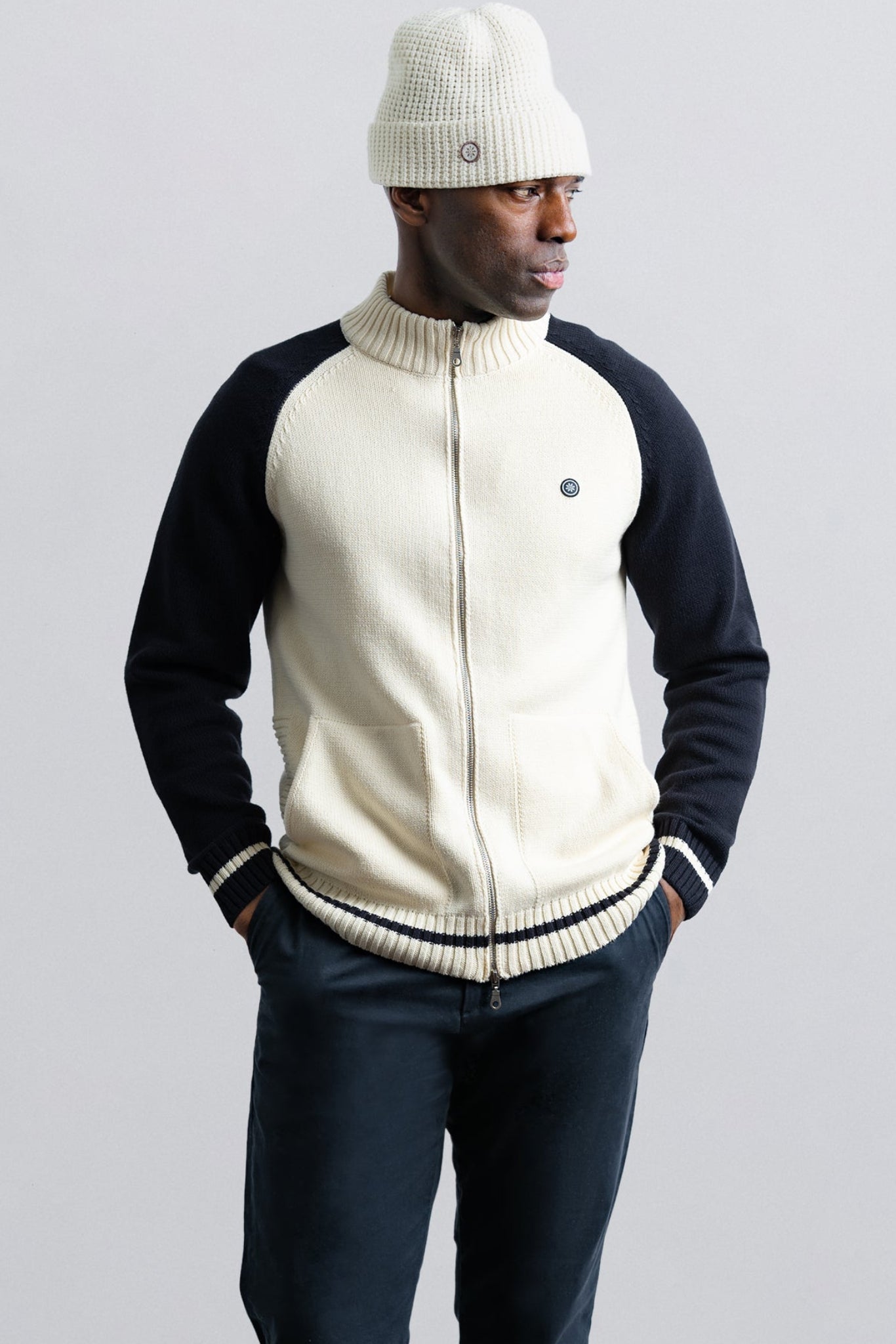 Man wearing a cream and navy varsity jacket with a white beanie on a light gray background