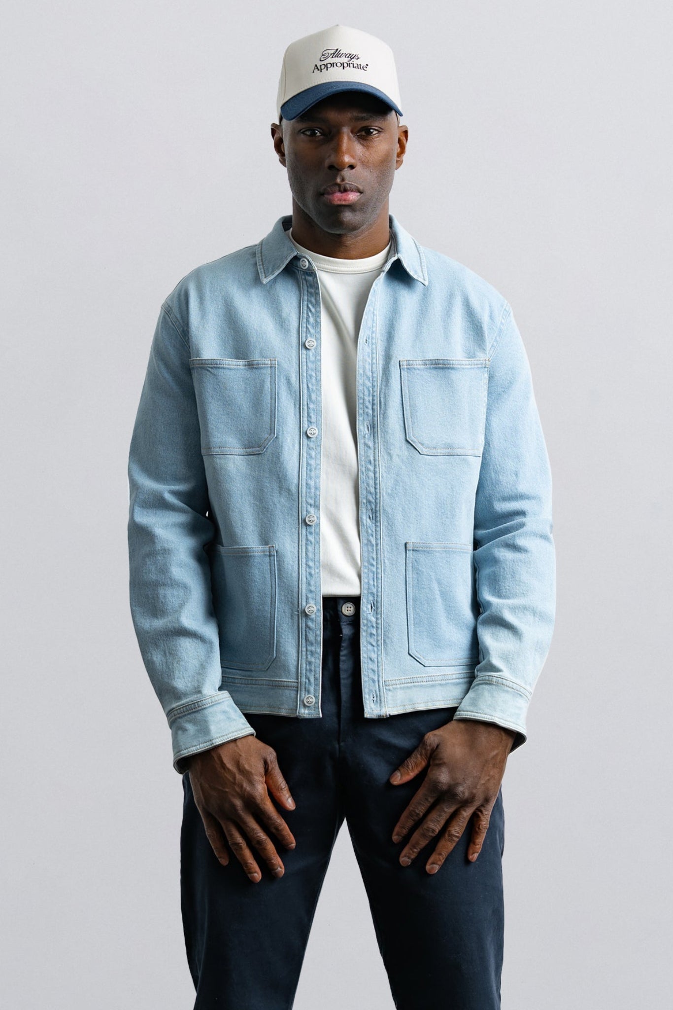 Man wearing a light blue denim jacket, white shirt, dark pants, and a white cap on a gray background
