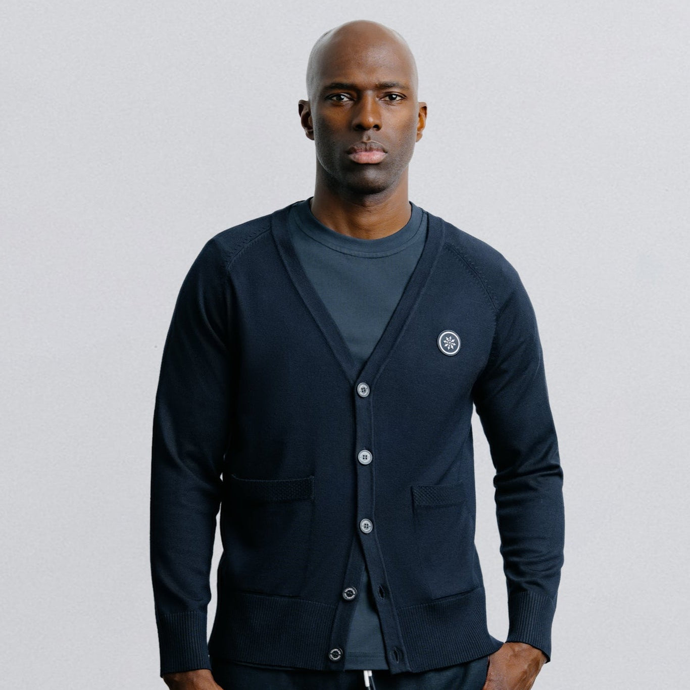 Man wearing a navy cardigan over a gray shirt with dark shorts on a light gray background