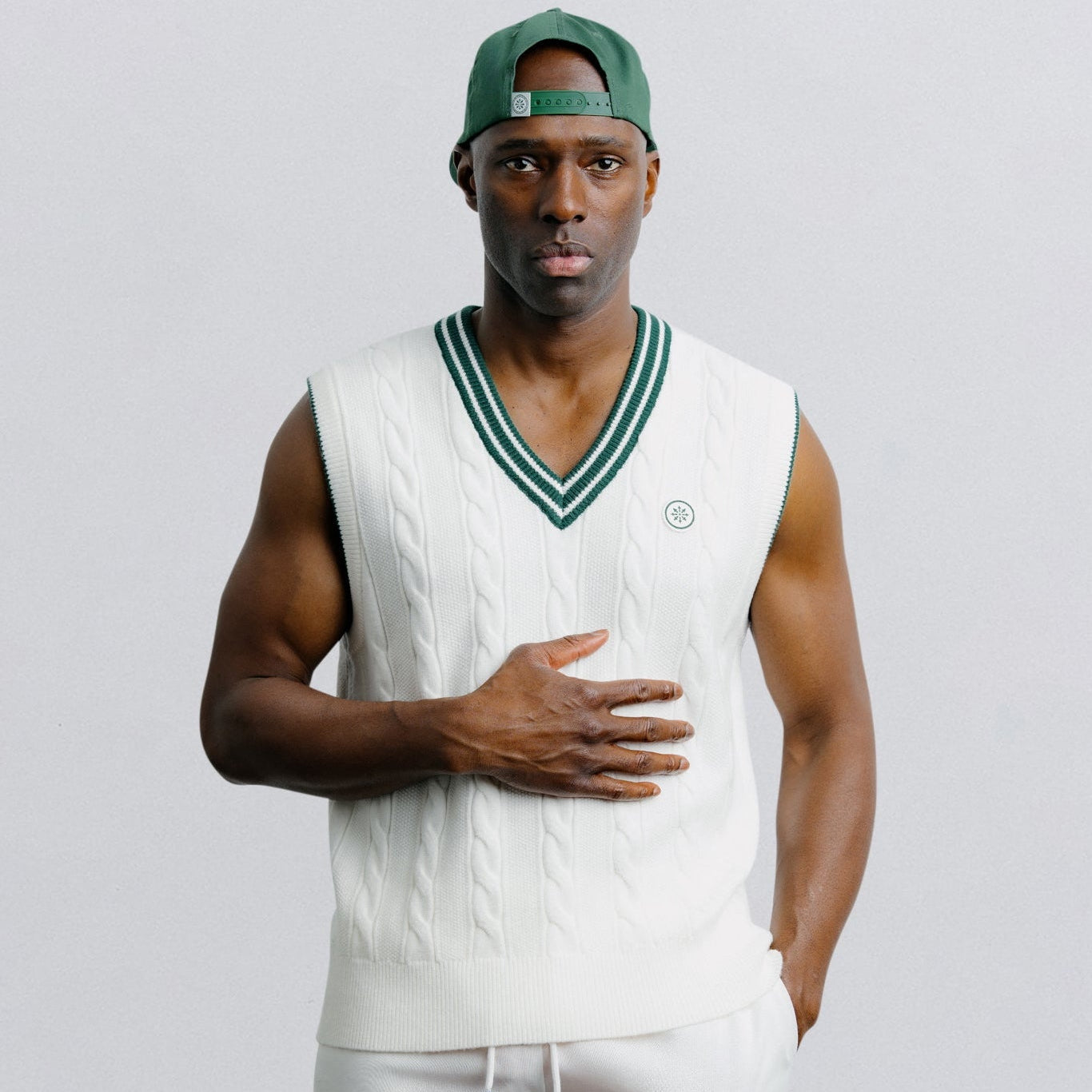 Man wearing a sleeveless shirt and shorts with a green cap on a light gray background