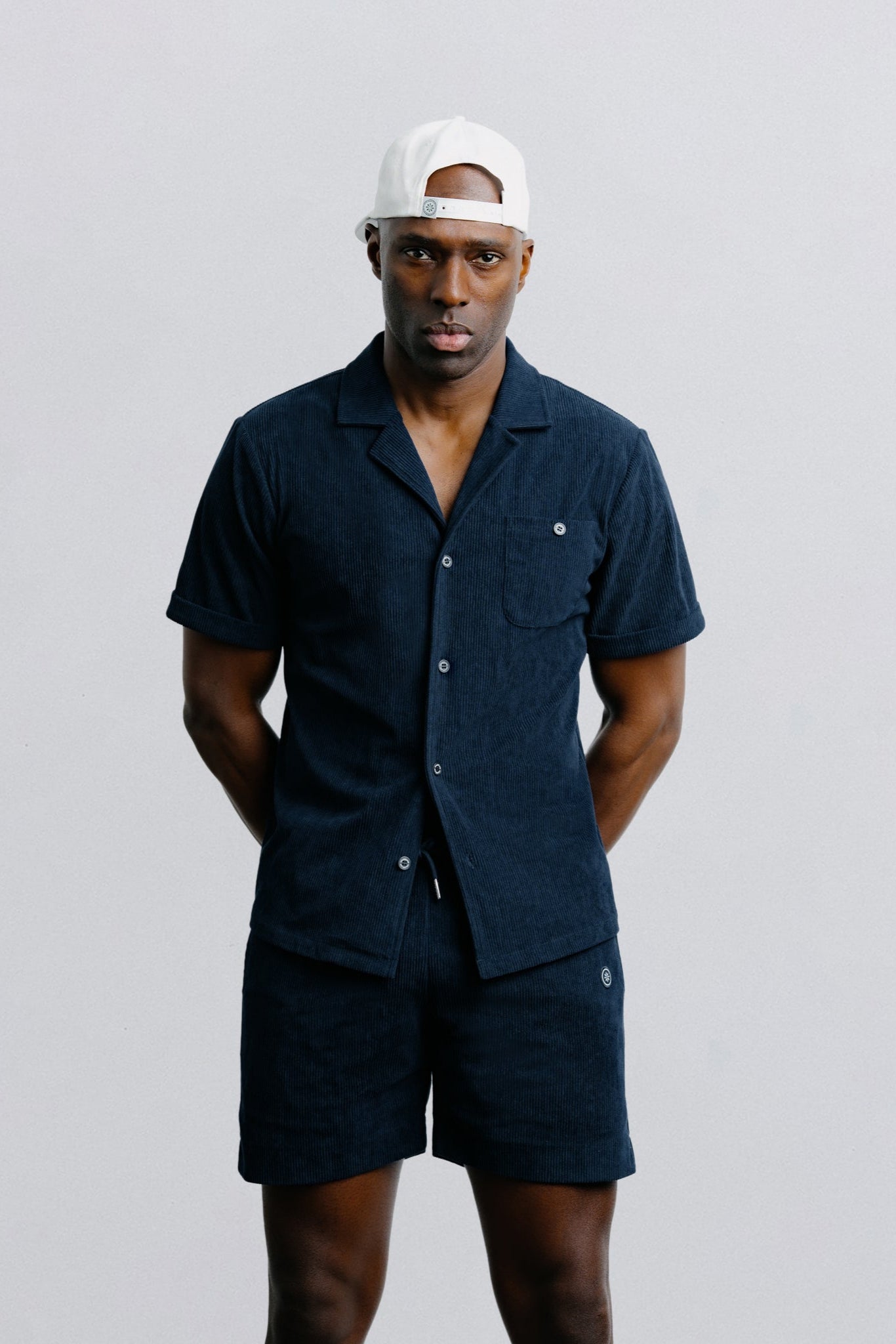 Man wearing a navy shirt and shorts set with a white cap on a light gray background