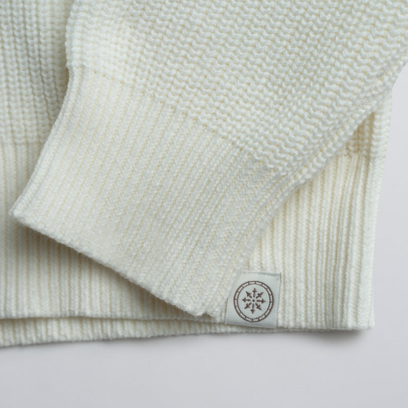 Close-up of a folded cream sweater with a logo on a plain background.