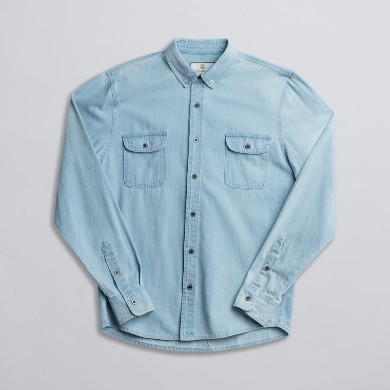 A blue denim shirt with two front pockets, button-up front, and long sleeves laid out flat on a white background.