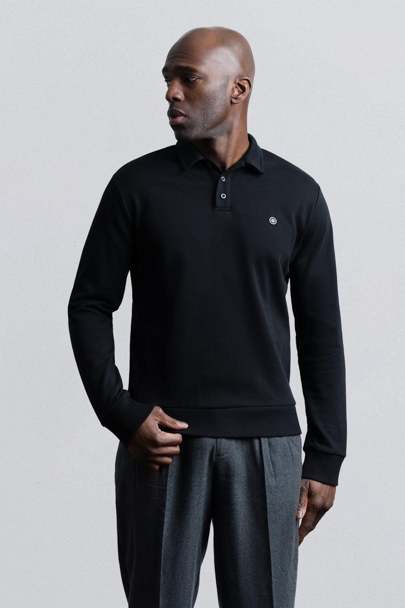 A man wearing a black long-sleeve polo shirt with a small embroidered logo on the chest, paired with dark gray trousers, standing against a light gray studio background.