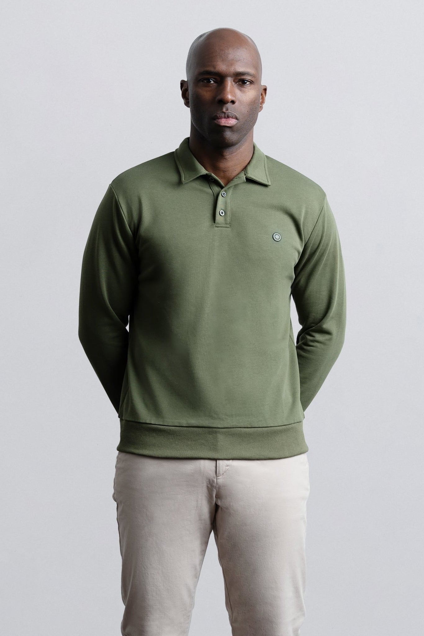 A man wearing an olive green long-sleeve polo shirt and beige trousers, standing against a light gray studio background.