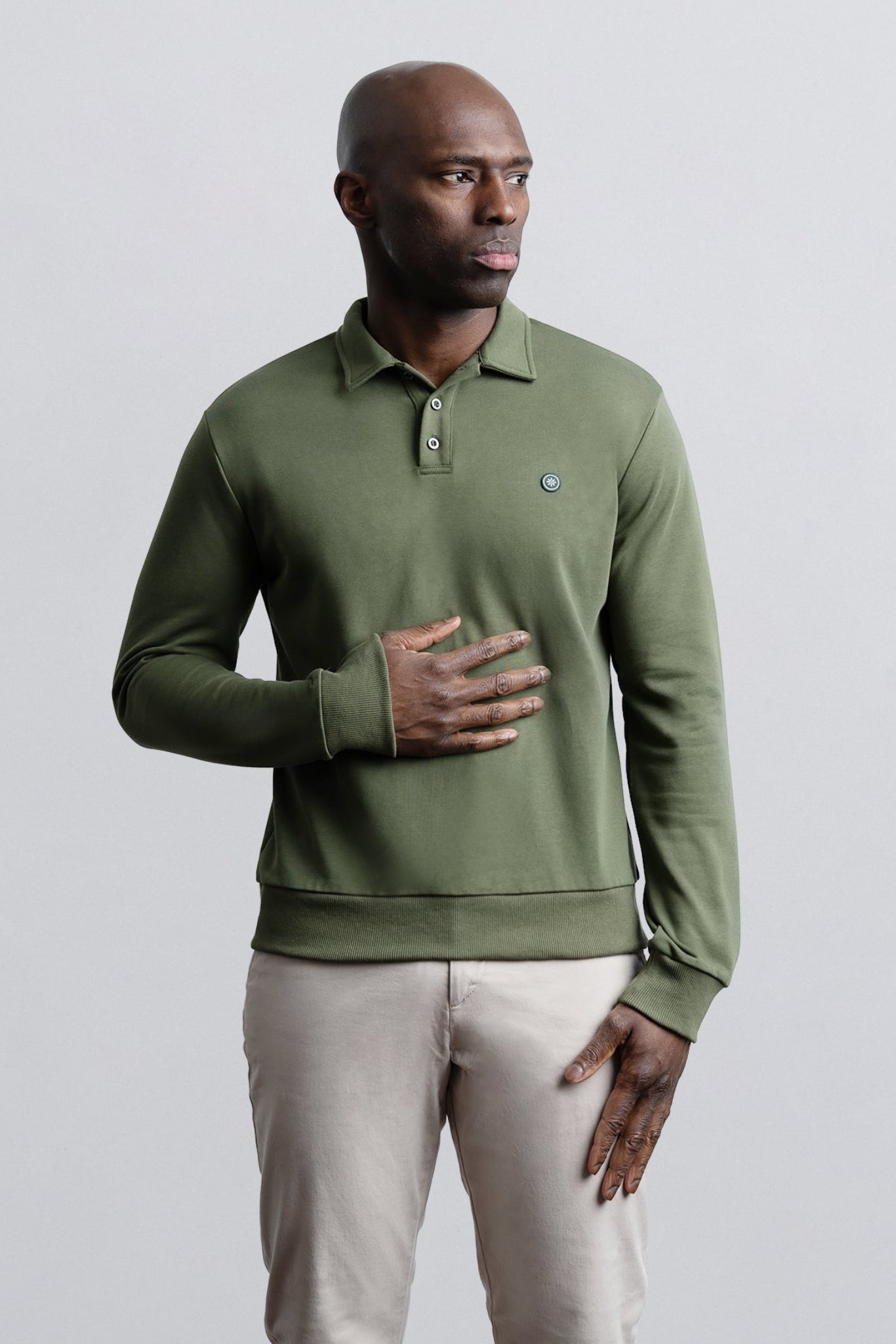 A man wearing an olive green long-sleeve polo shirt with a small embroidered crocodile logo on the chest, paired with light beige trousers, standing against a solid light gray background.