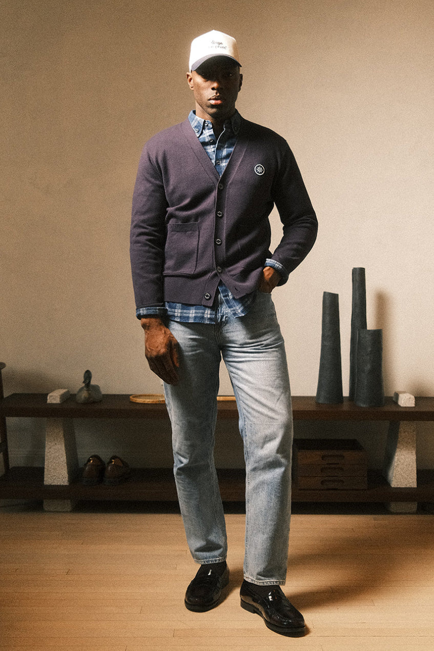 A man wearing a dark gray cardigan, light blue jeans, a blue plaid shirt, black loafers, and a white baseball cap stands in a minimalist room with a beige wall and wooden floor.