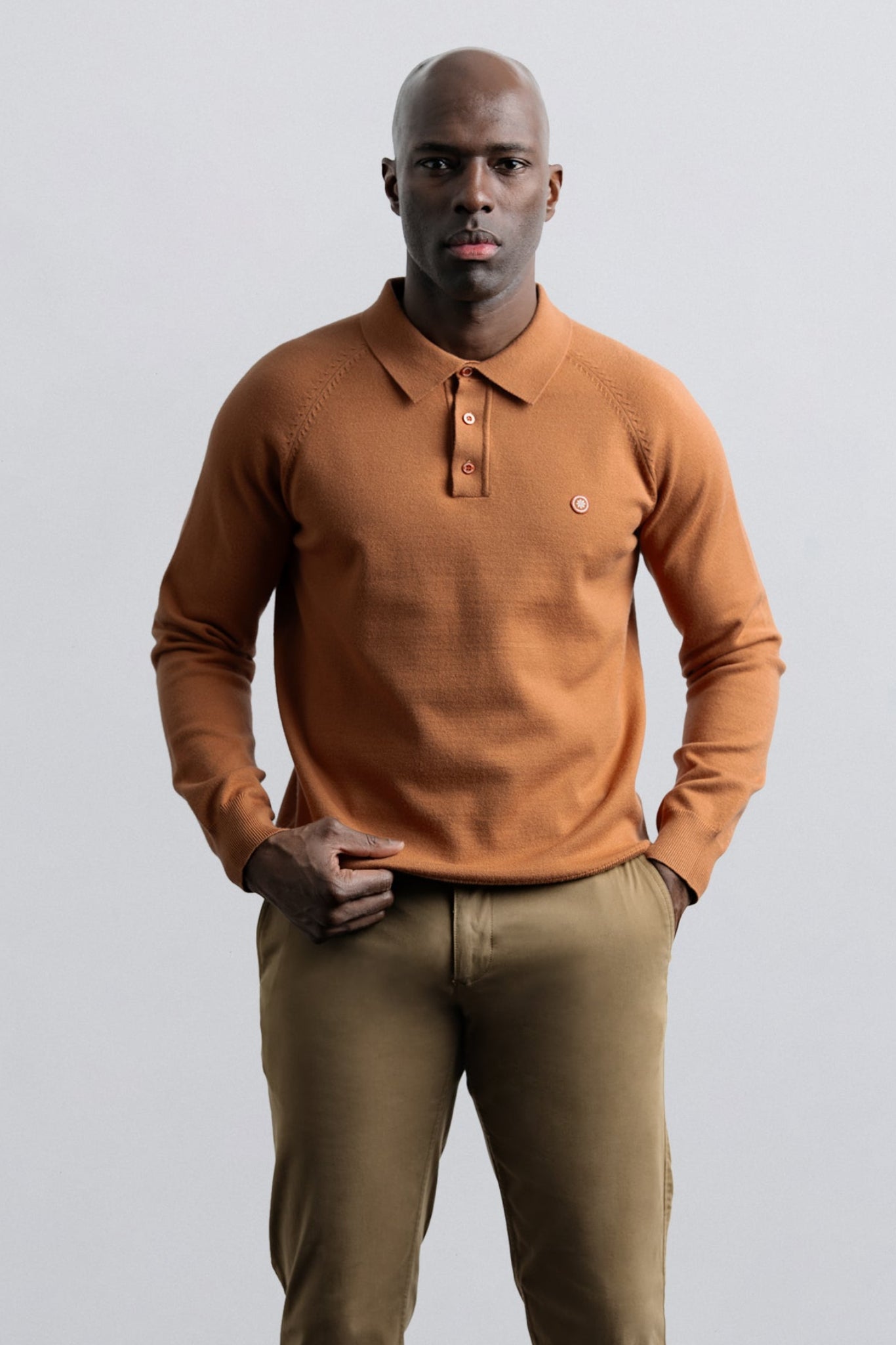 A man wearing a rust-orange knit polo sweater and khaki trousers, standing against a light gray studio background.