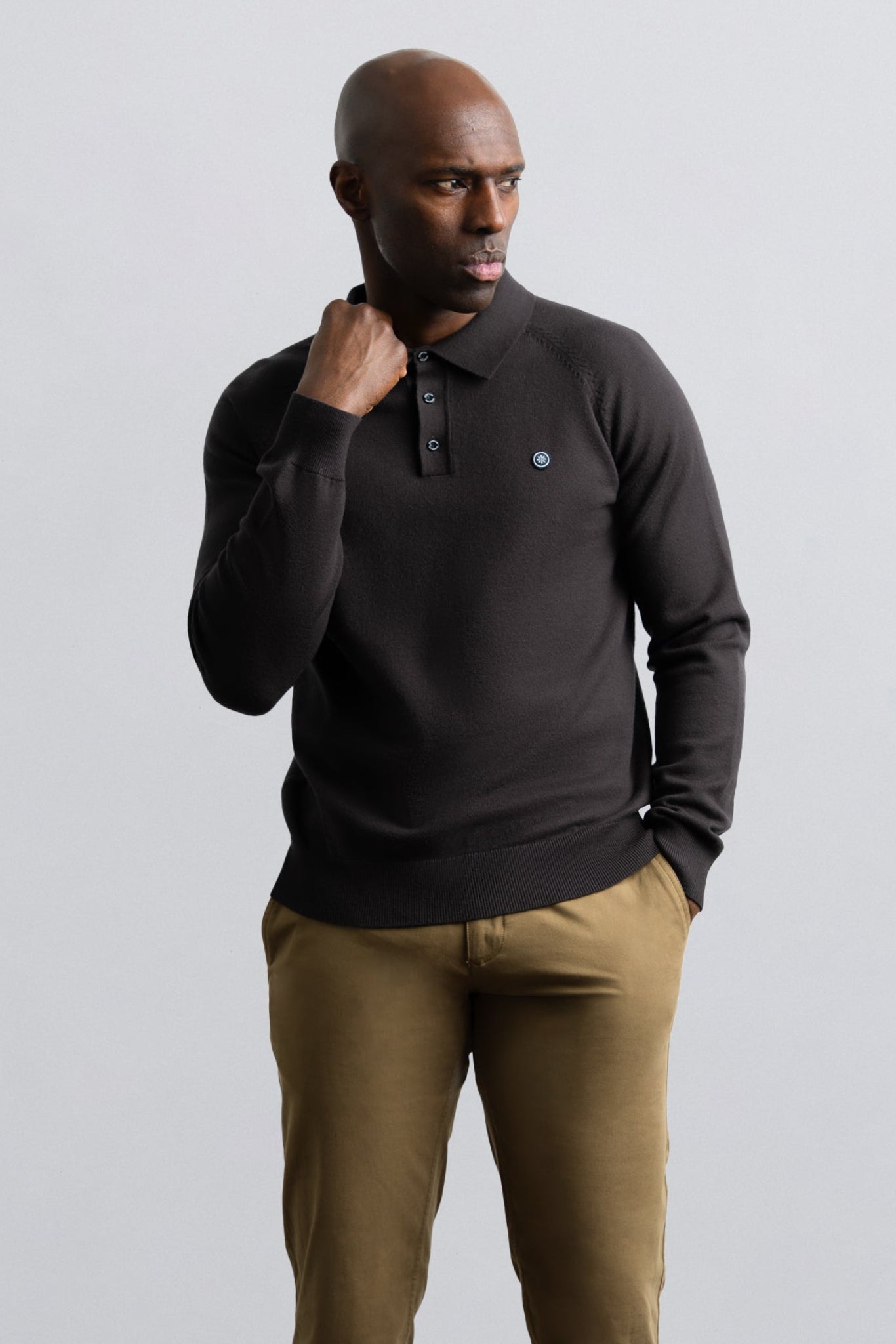A man wearing a dark charcoal long-sleeve polo shirt with a small blue embroidered logo on the chest, paired with khaki trousers, standing against a light gray studio background.