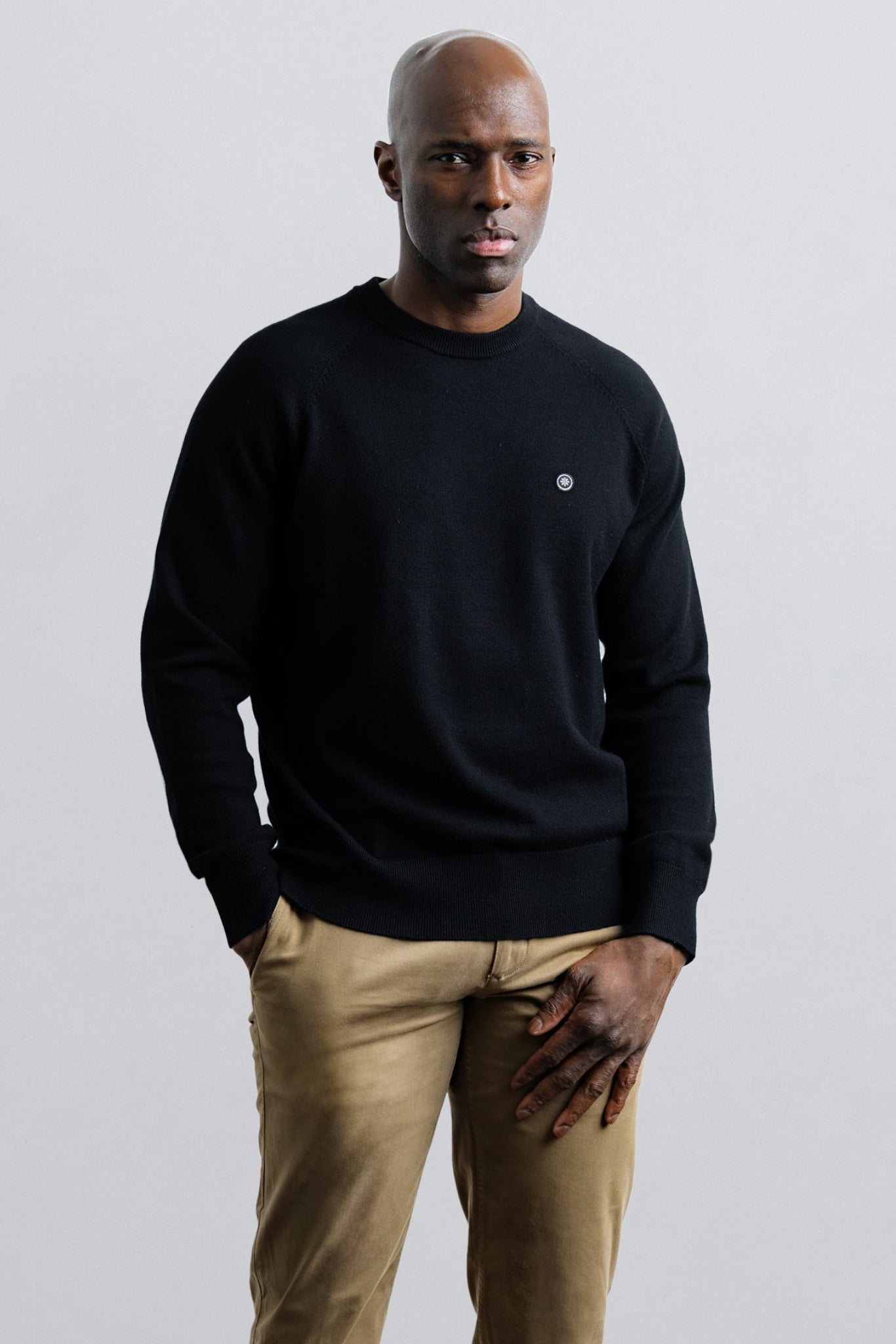 Man wearing a black sweater and beige pants against a plain background