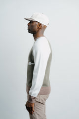 Man wearing a white cap, white long-sleeve shirt, and patterned sweater against a light gray background