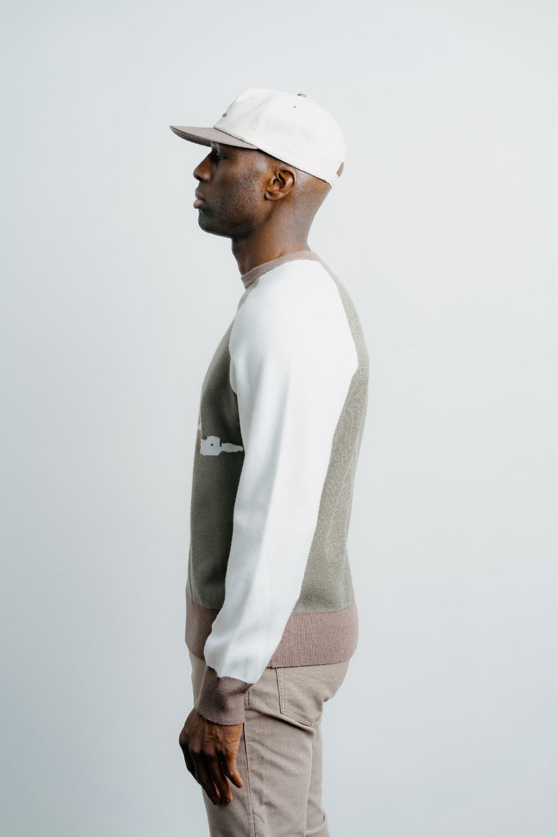 Man wearing a white cap, white long-sleeve shirt, and patterned sweater against a light gray background