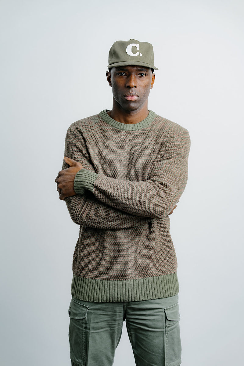 Man wearing a brown sweater and green cap with a 'C' logo on a light gray background