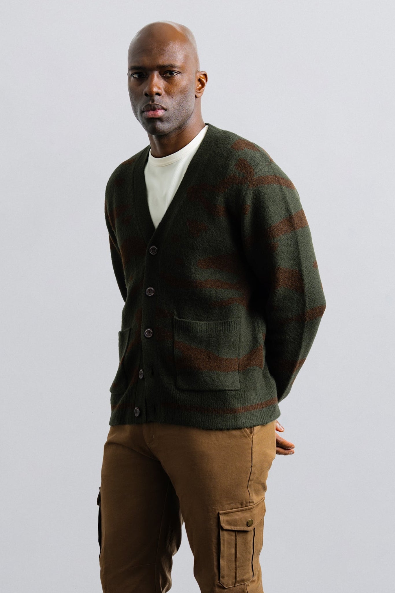 Man wearing a green cardigan with brown stripes and brown pants on a light gray background