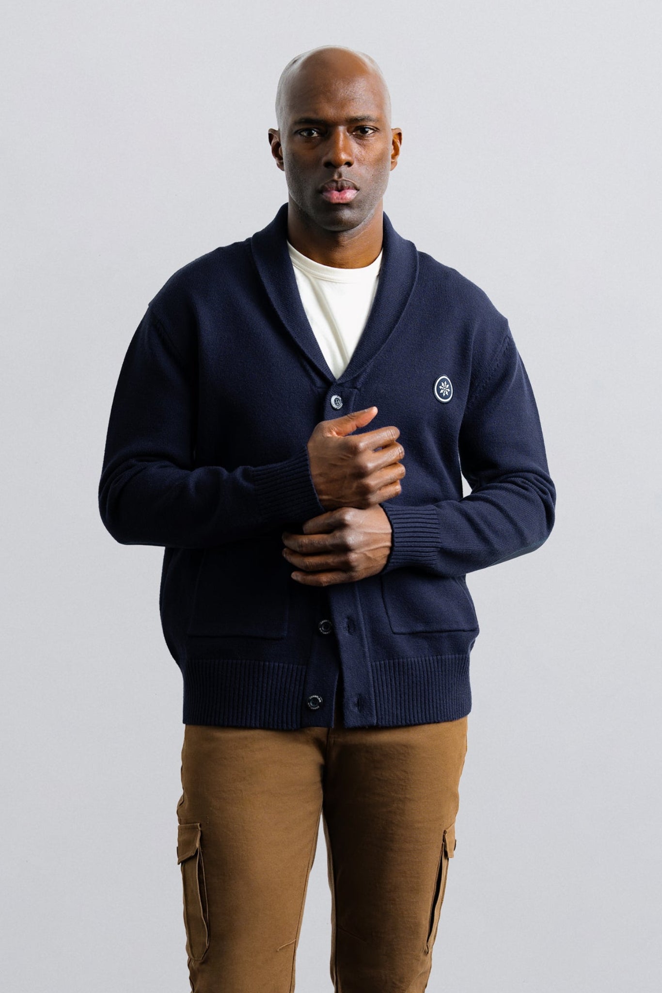 Man wearing a navy blue cardigan over a white shirt with brown pants on a light gray background