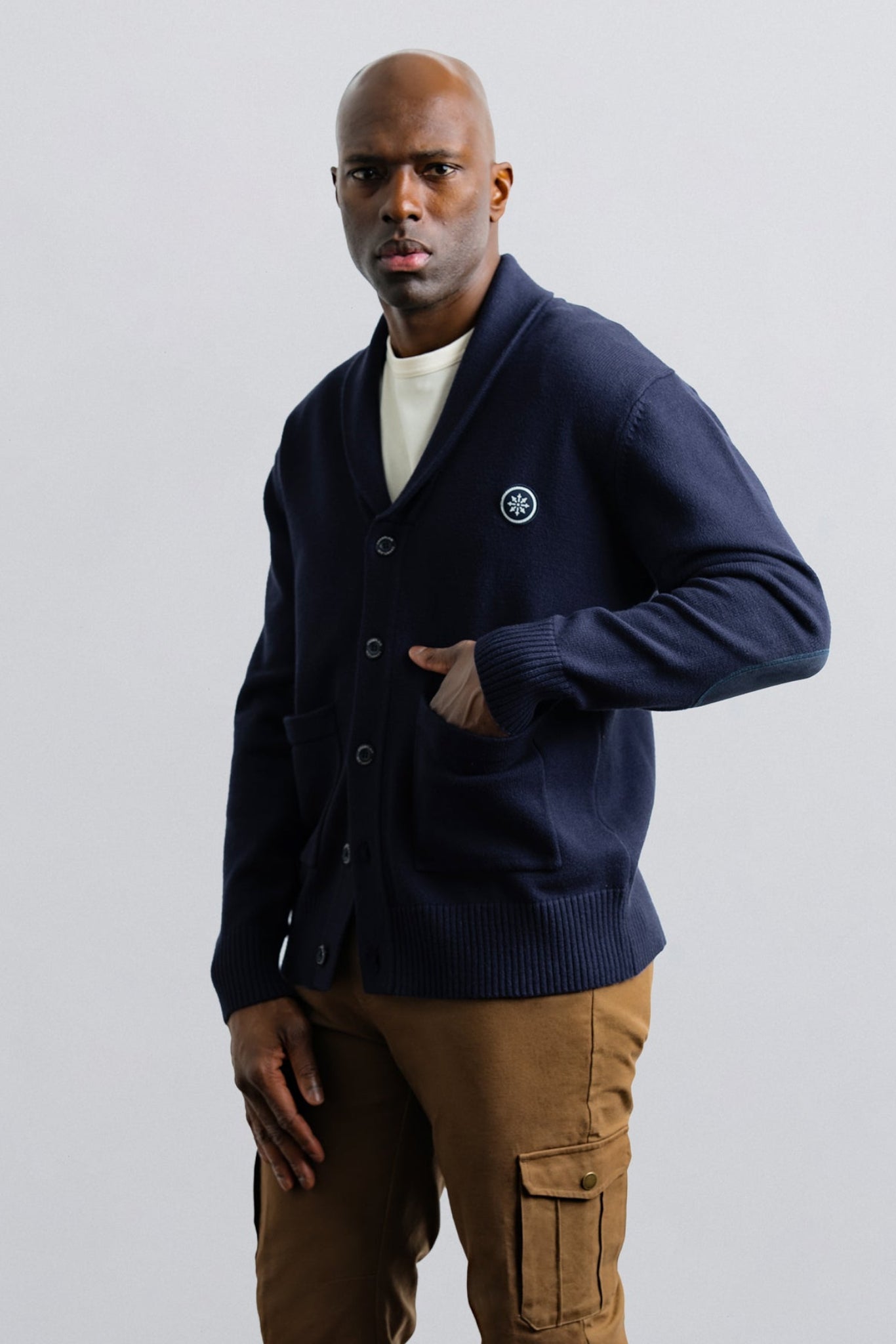 Man wearing a navy cardigan over a white shirt with brown pants on a light gray background