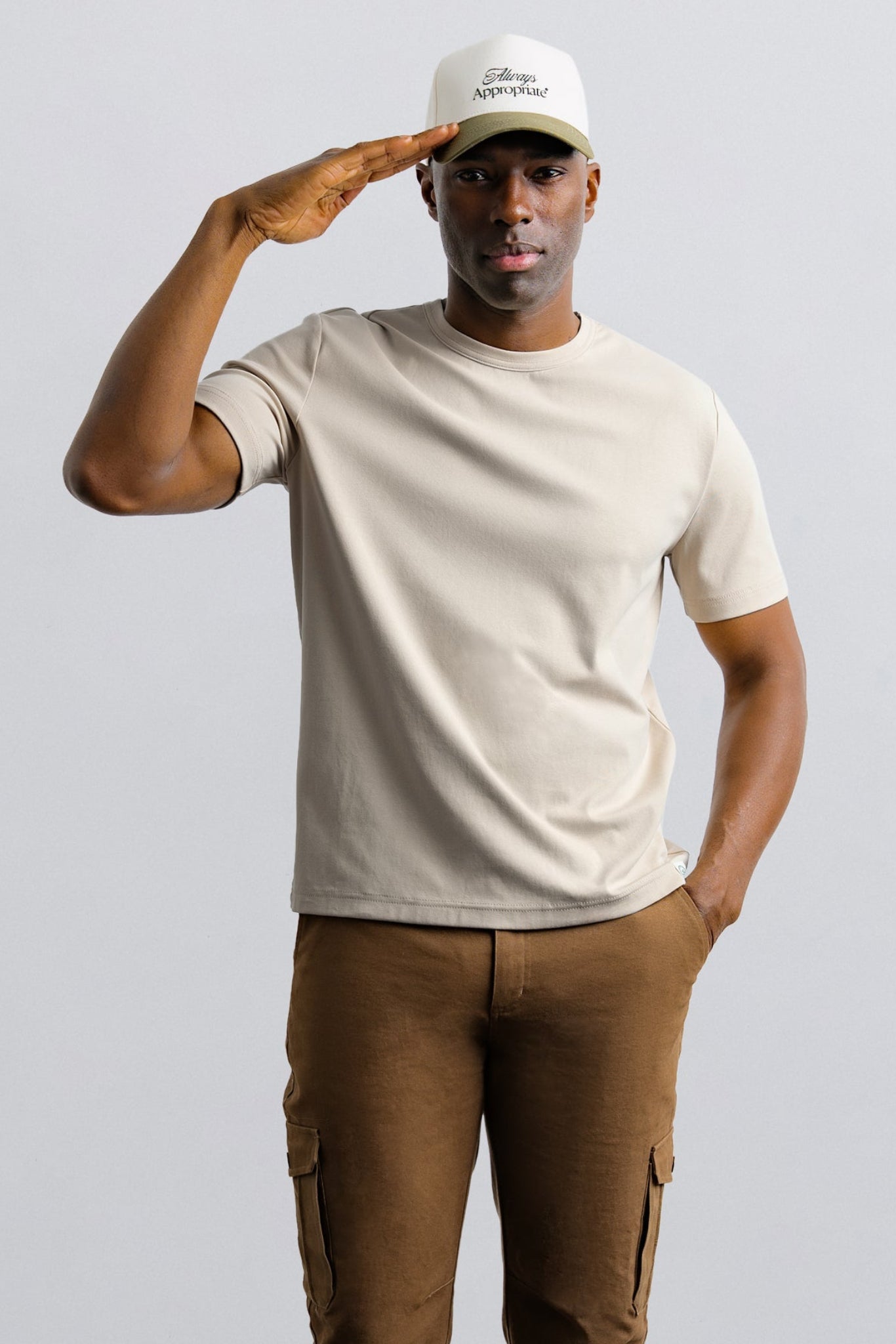 Man wearing a beige t-shirt and brown pants saluting with a white cap on a light gray background