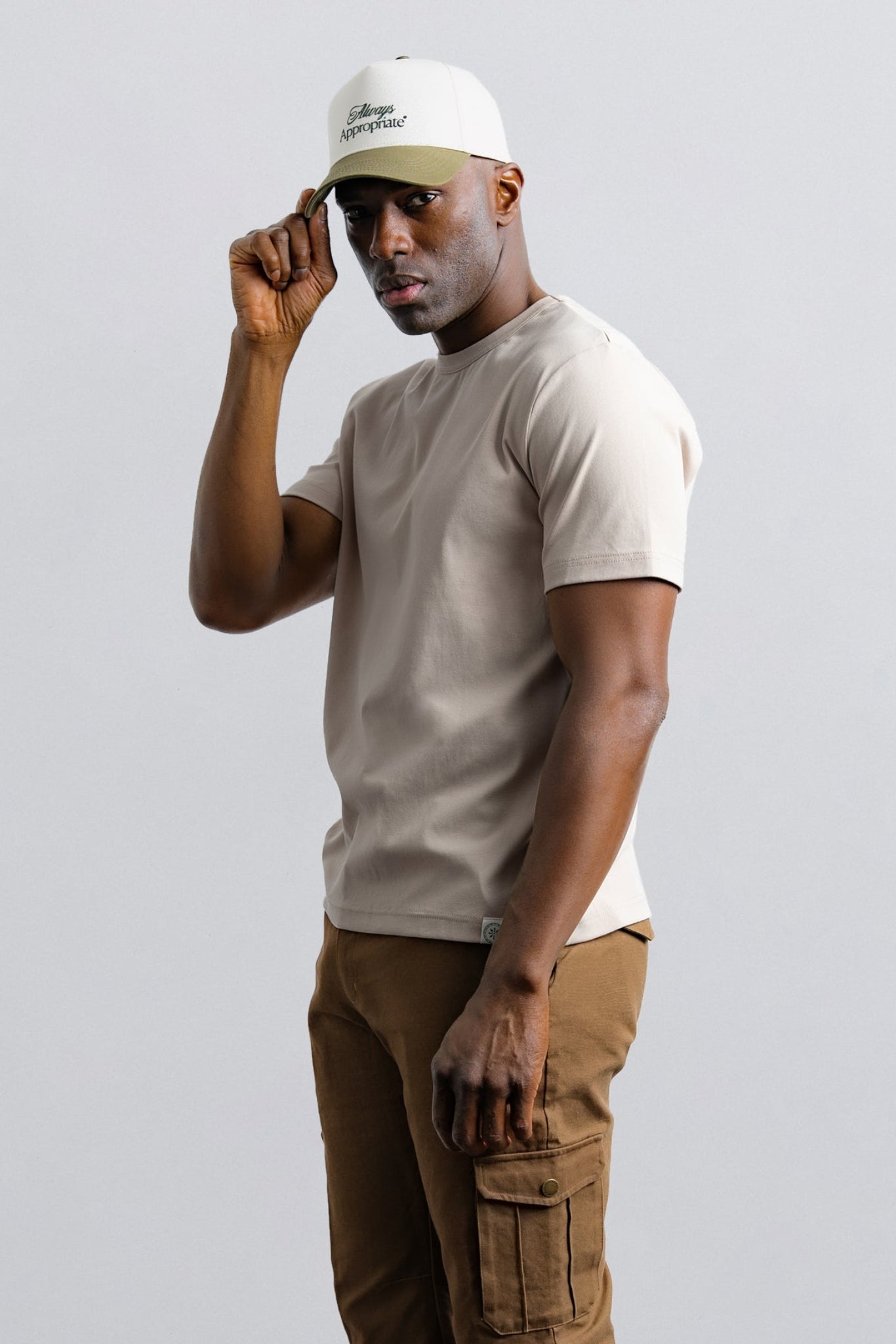 Man wearing a beige t-shirt and brown pants with a white cap on a light gray background