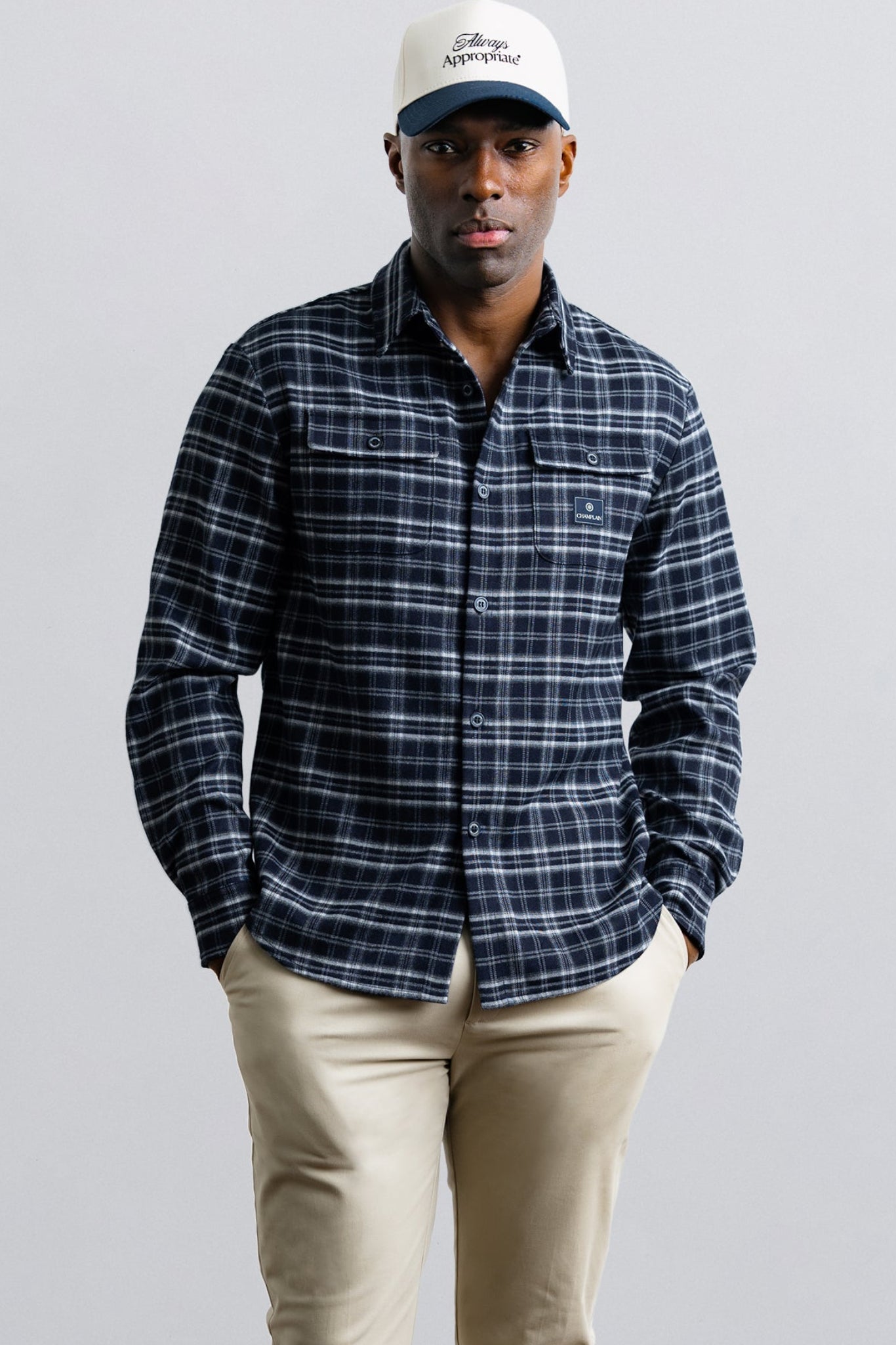 Man wearing a blue plaid shirt, beige pants, and a white cap with a logo on a gray background