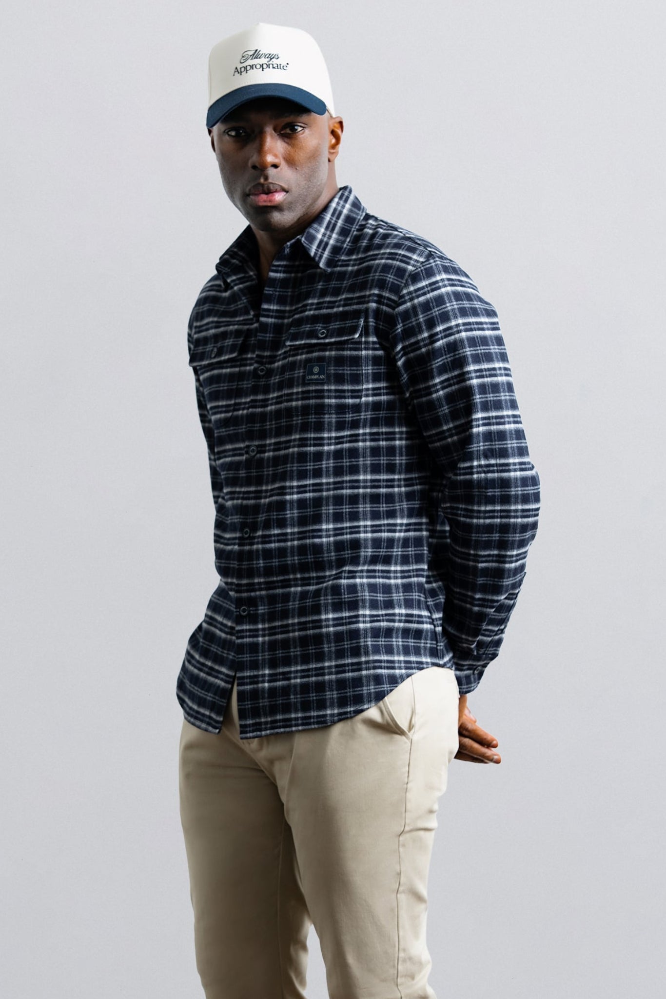 Man wearing a plaid shirt, beige pants, and a cap on a gray background