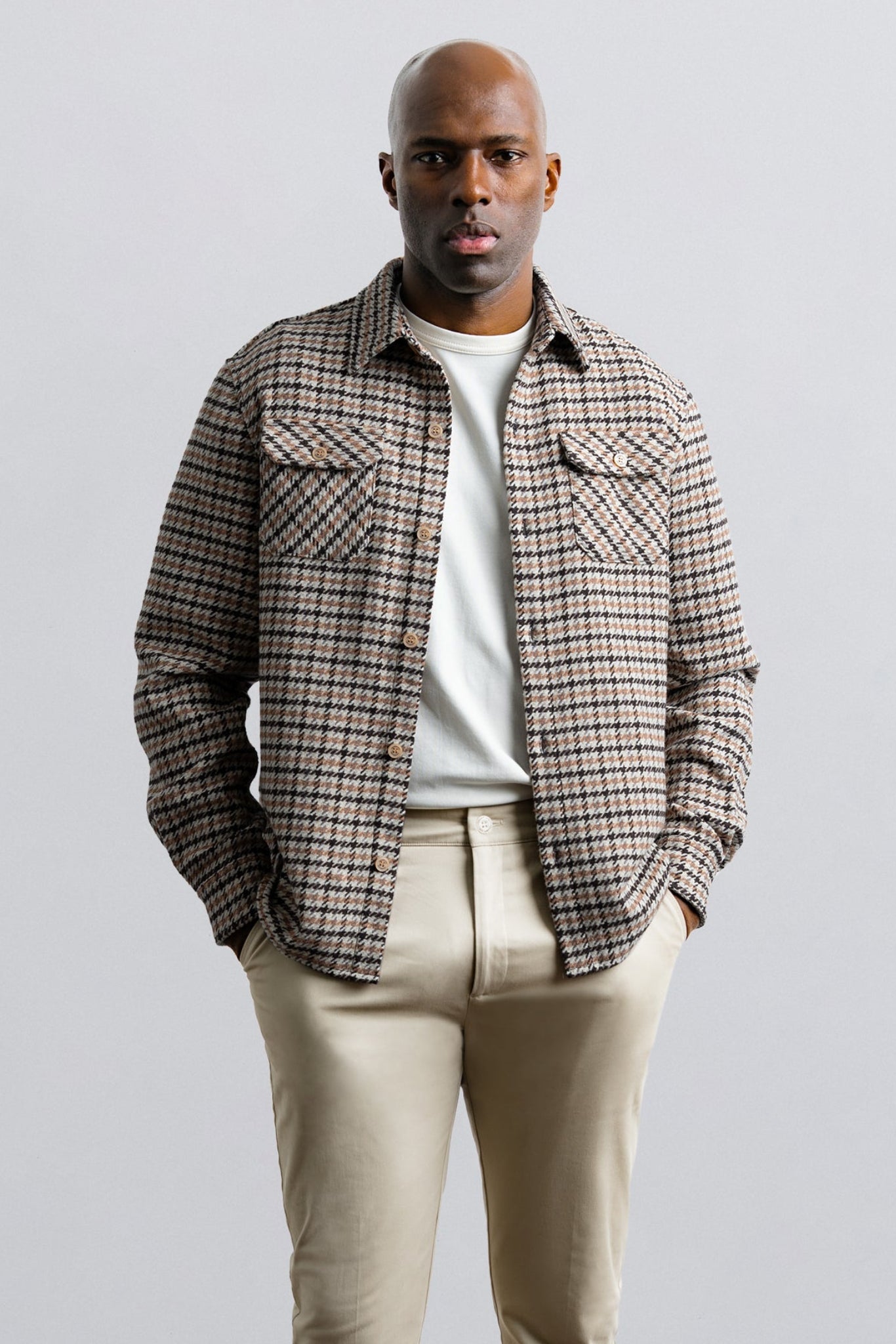 Man wearing a plaid shirt over a white t-shirt with beige pants on a light gray background