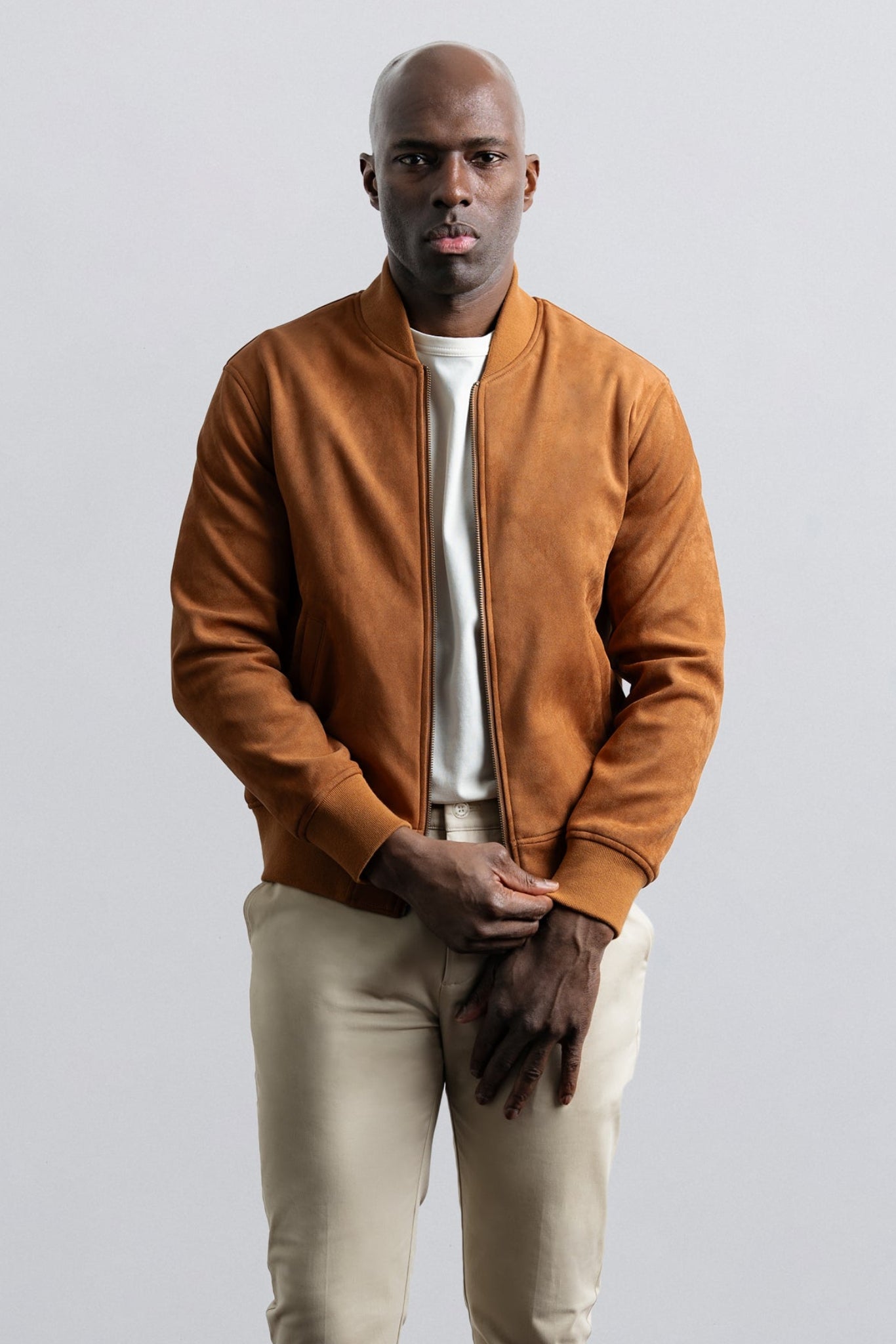 Man wearing a brown jacket over a white shirt and beige pants on a light gray background