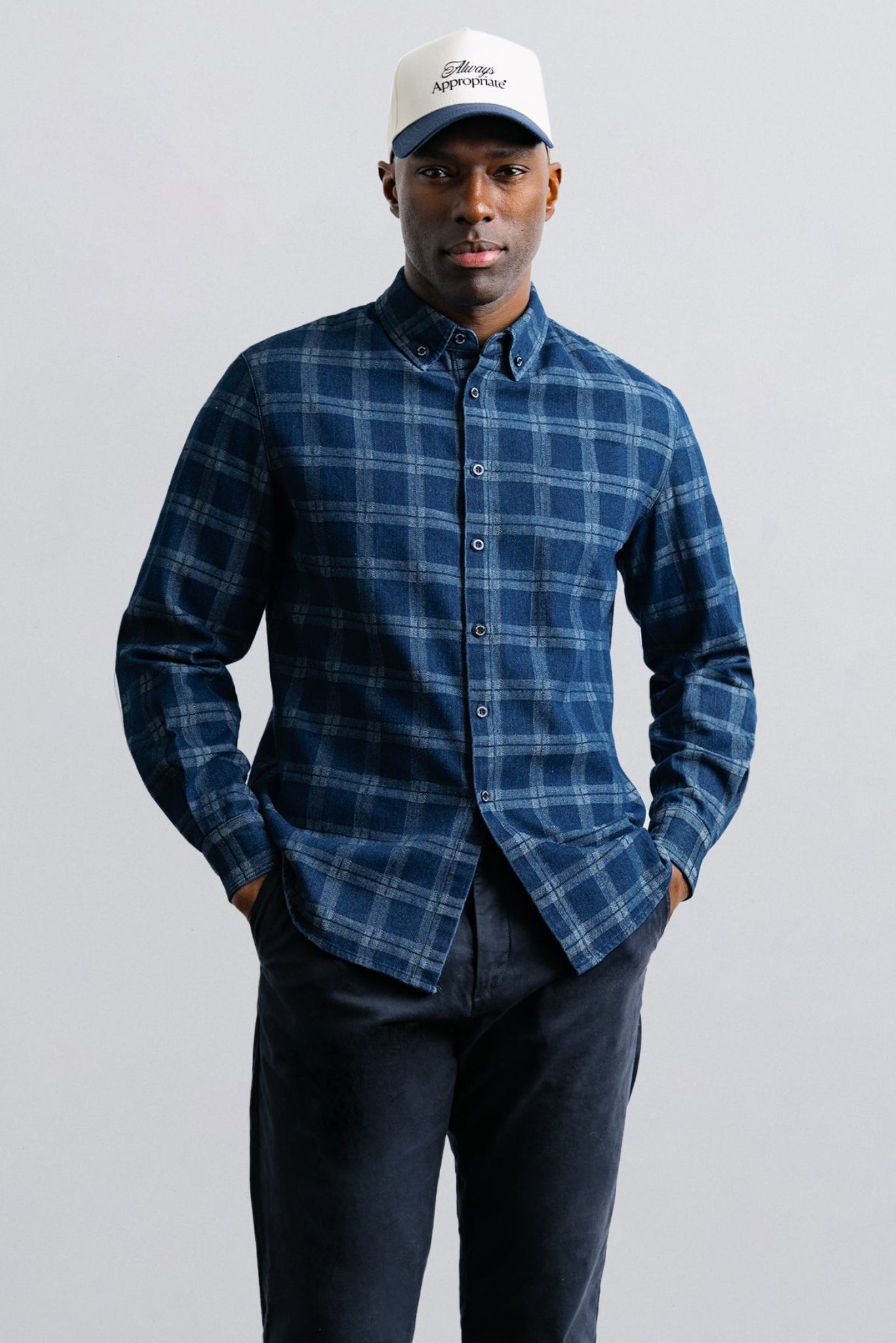Man wearing a blue plaid shirt and white cap against a gray background