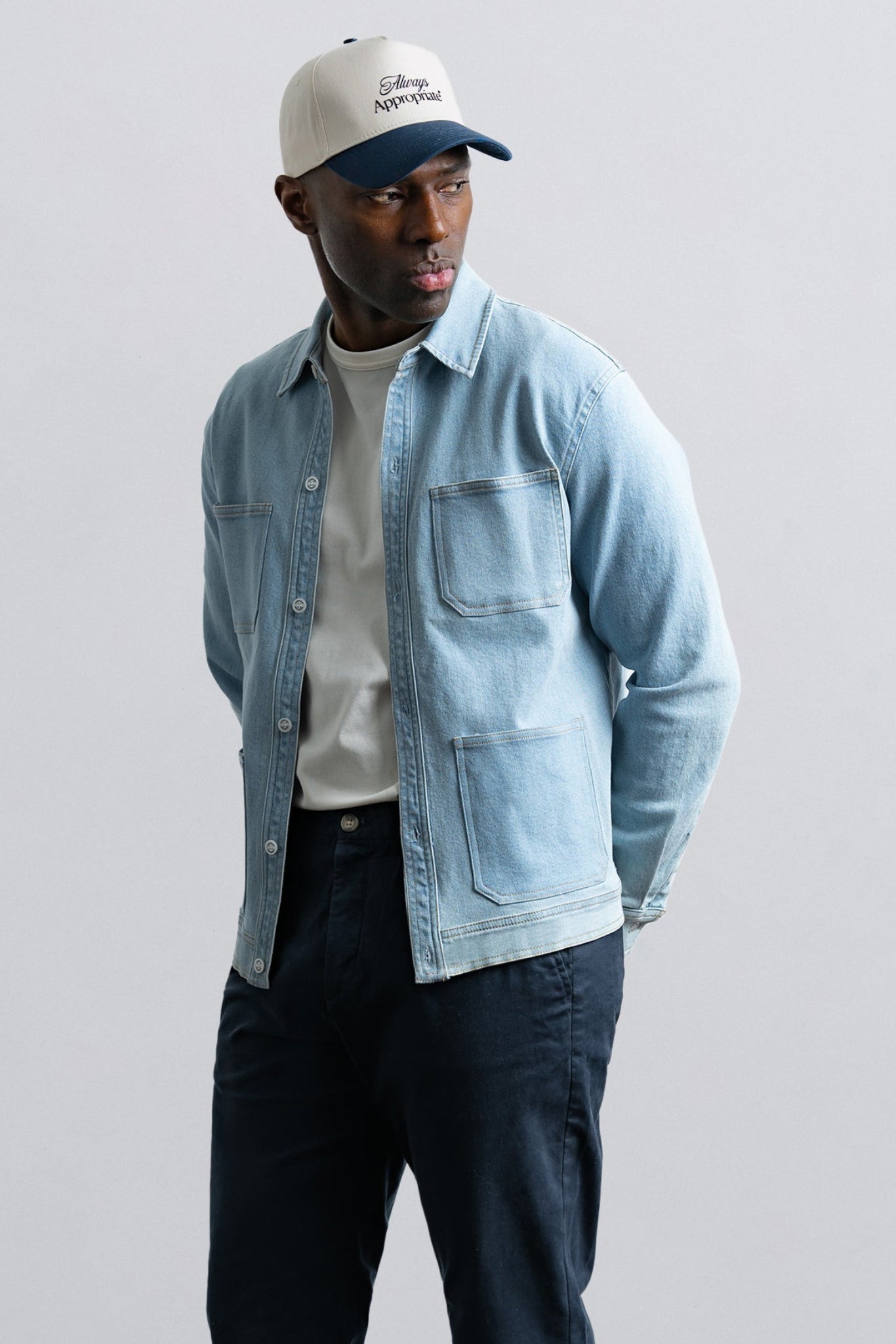 Man wearing a light blue denim jacket, white t-shirt, dark pants, and a cap on a plain background