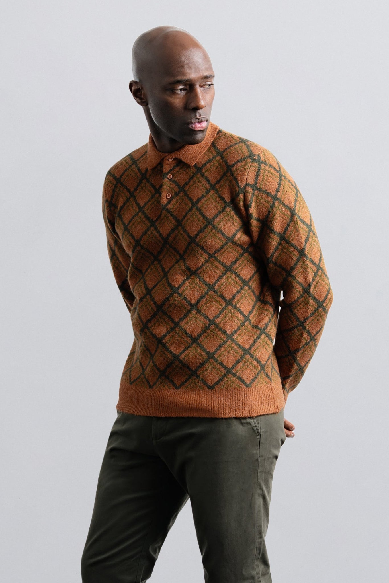 Man wearing a patterned brown sweater and green pants against a light gray background