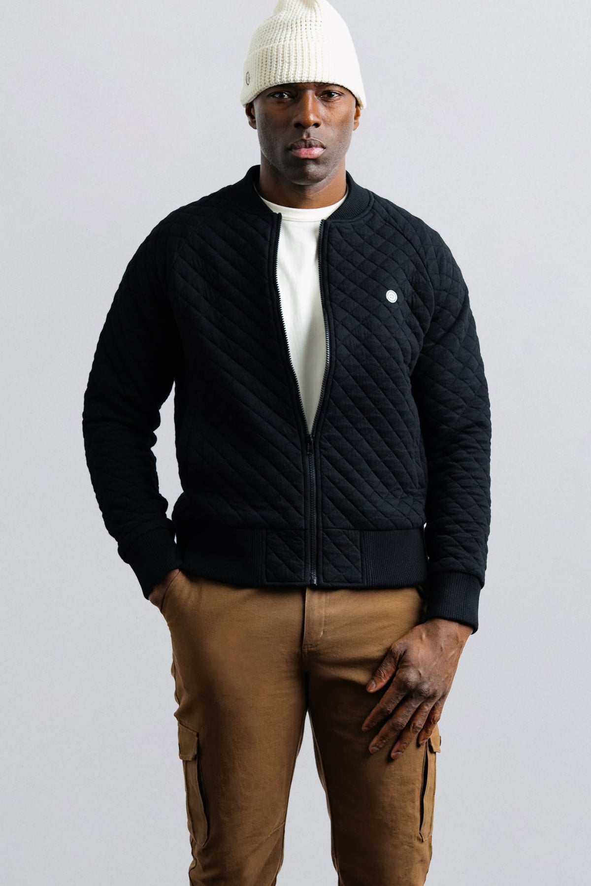 Man wearing a black quilted jacket, white beanie, and brown pants on a light gray background