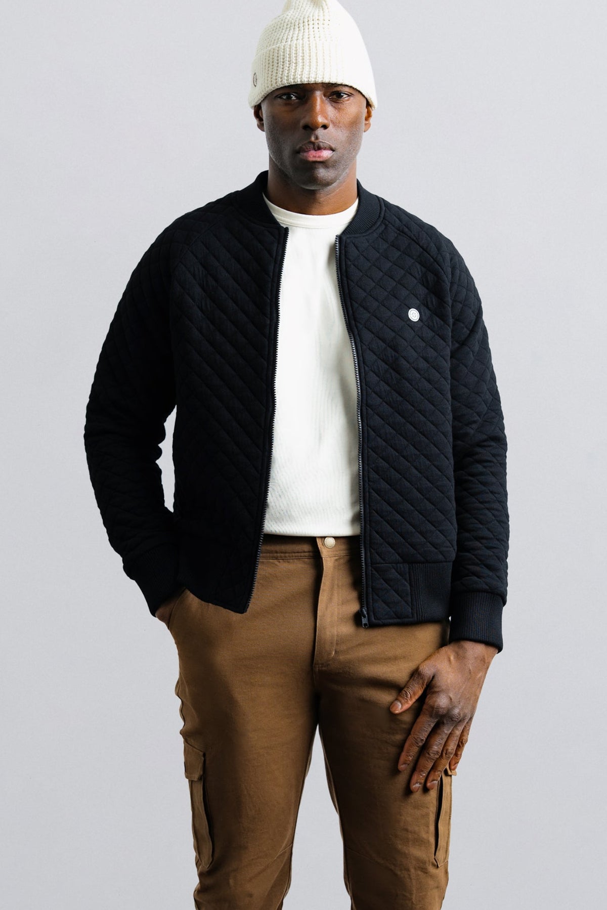 Man wearing a black quilted jacket, white beanie, and brown pants on a light gray background