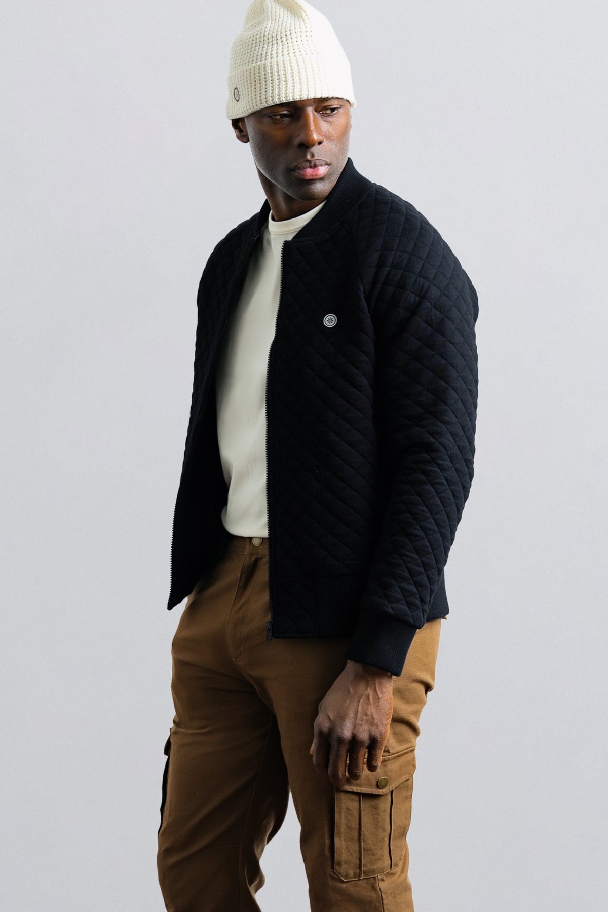Man wearing a black quilted jacket, white beanie, and brown pants on a light gray background
