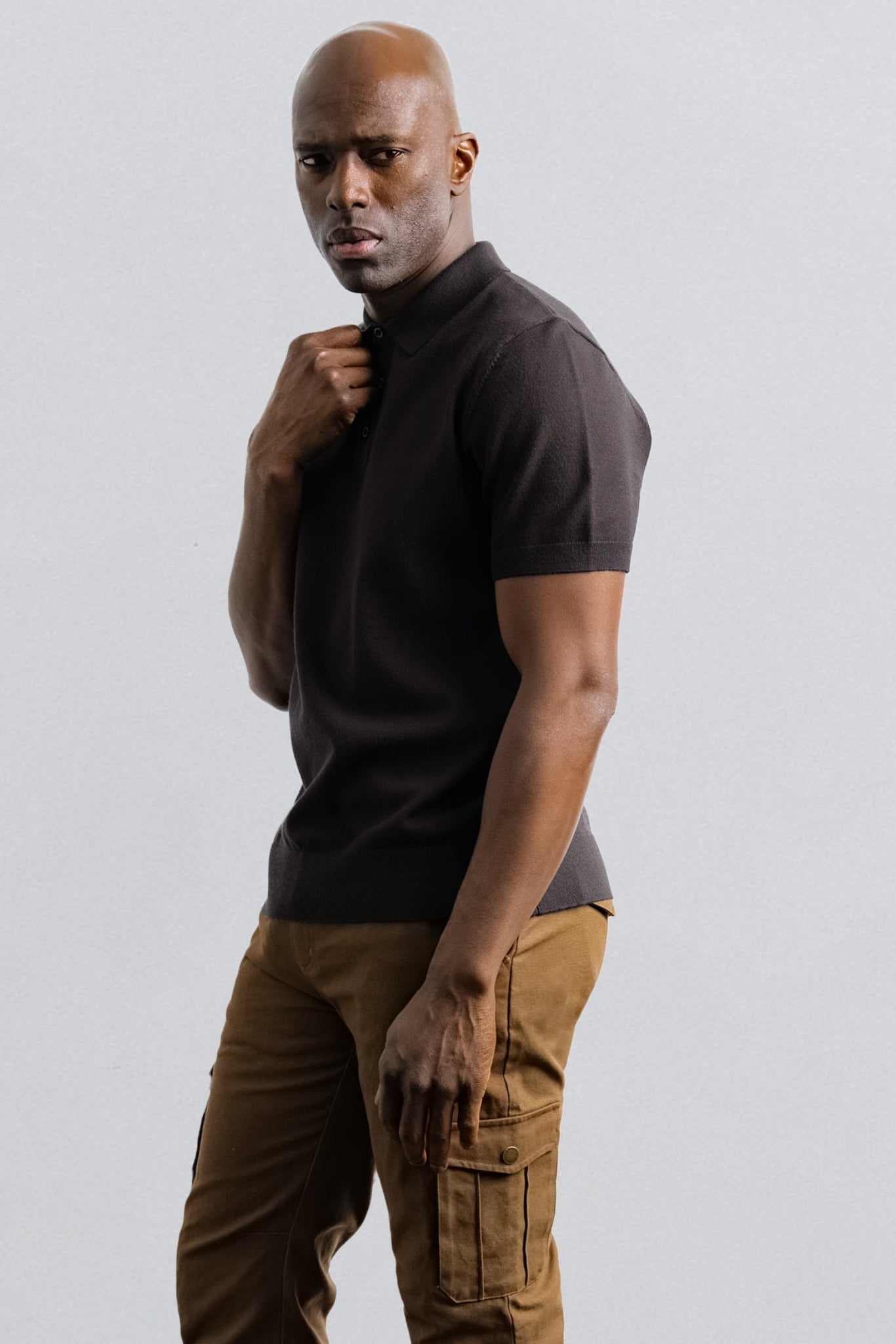 A man wearing a dark brown short-sleeve Polo knit shirt and brown cargo pants, standing against a light gray studio background.