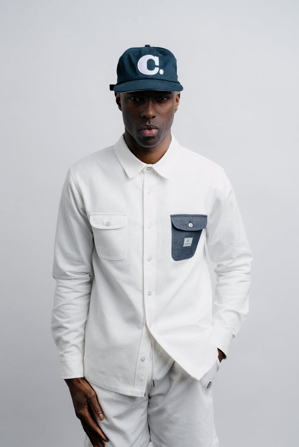 Model wearing a white denim overshirt with a blue champlain C hat.