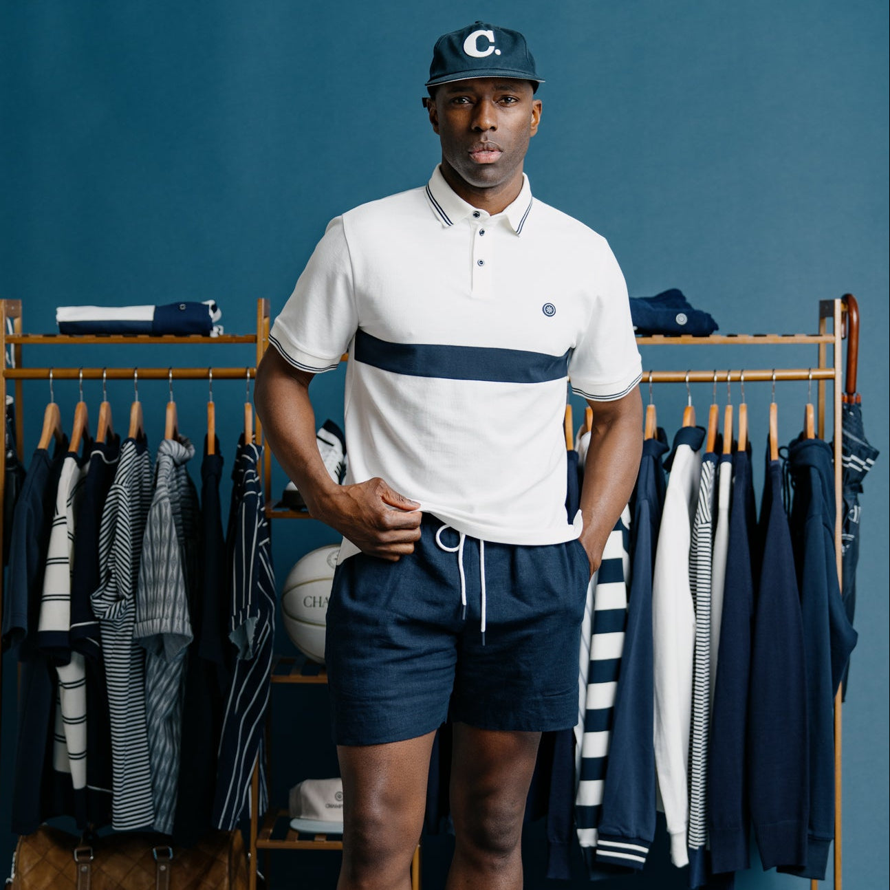 Man wearing a white polo shirt with a navy stripe and dark shorts, standing in front of a clothing rack.
